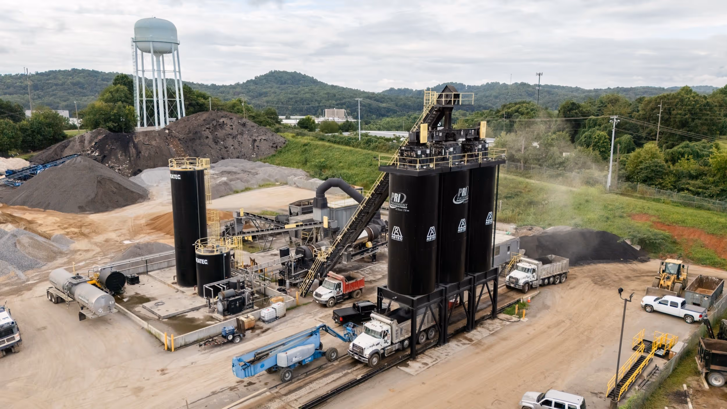Industrial asphalt production facility with large black storage silos, several trucks, and construction equipment surrounded by mounds of gravel and a water tower in the background.