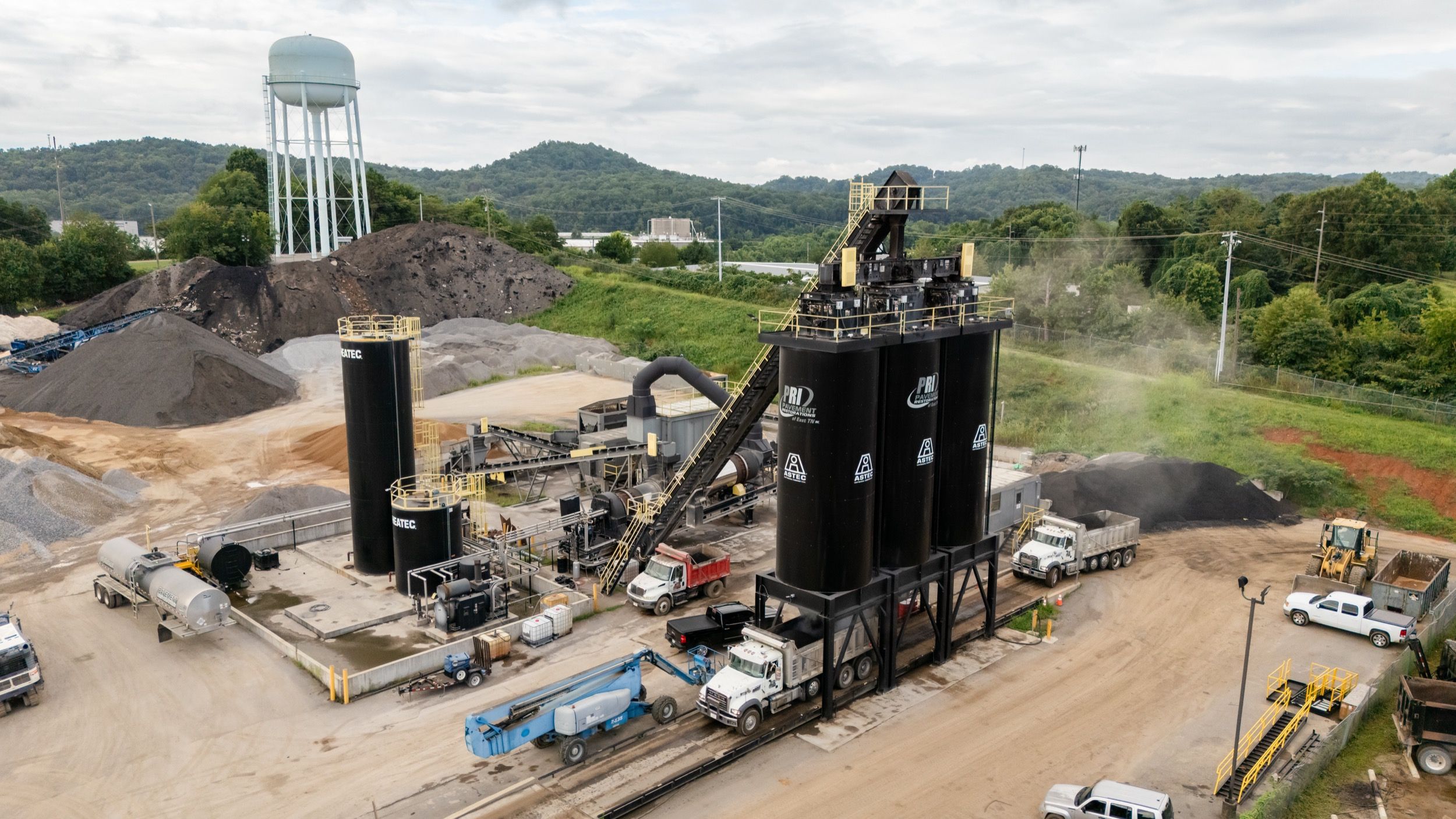 Industrial asphalt production facility with large black storage silos, several trucks, and construction equipment surrounded by mounds of gravel and a water tower in the background.
