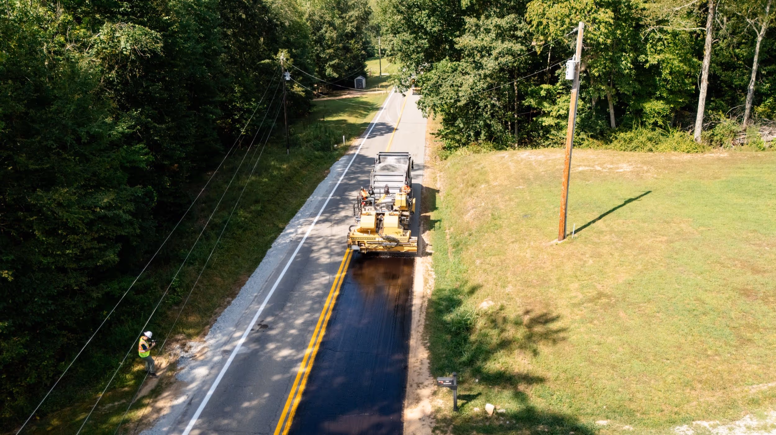 A machine applying fresh asphalt on a rural two-lane road surrounded by trees and grass, with a worker standing nearby.