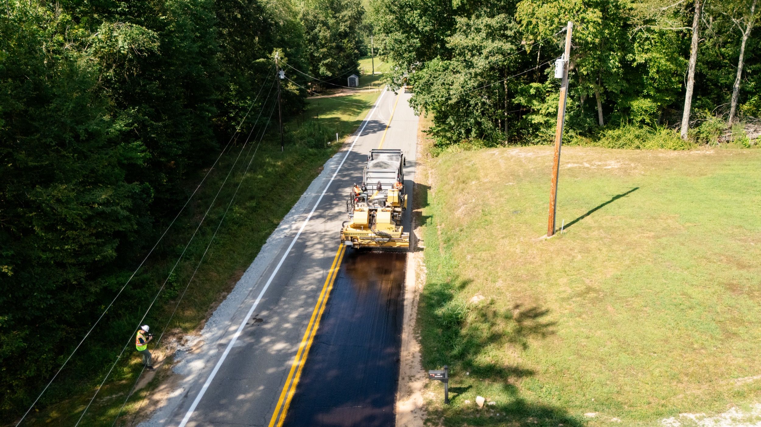 A machine applying fresh asphalt on a rural two-lane road surrounded by trees and grass, with a worker standing nearby.