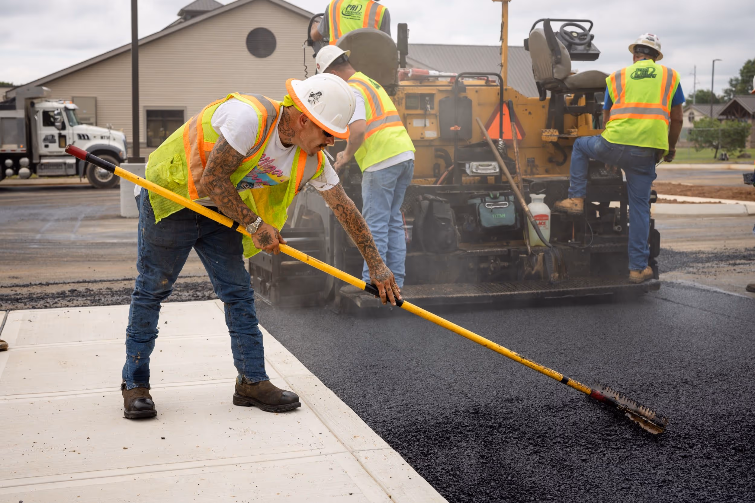 Construction workers wearing safety vests and hard hats paving a road with asphalt using a broom and paving machine.