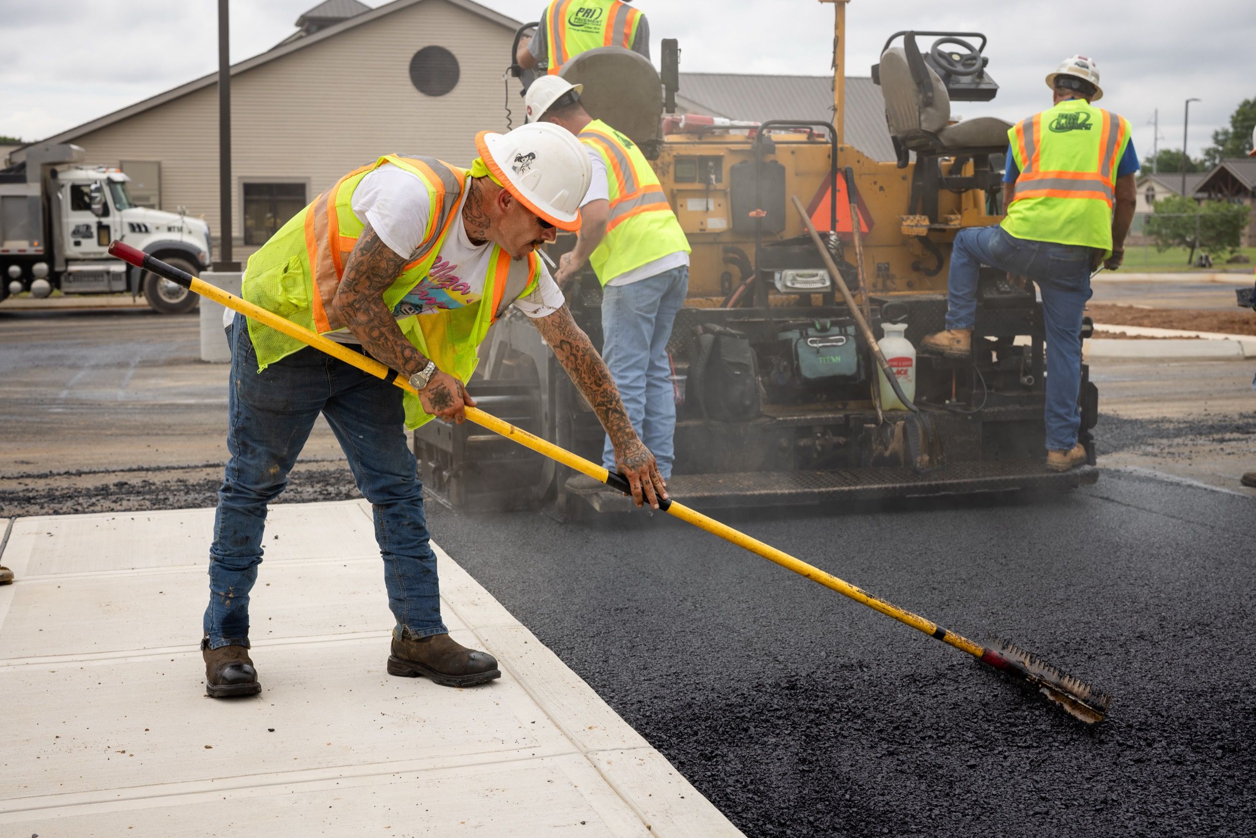 Construction workers wearing safety vests and hard hats paving a road with asphalt using a broom and paving machine.