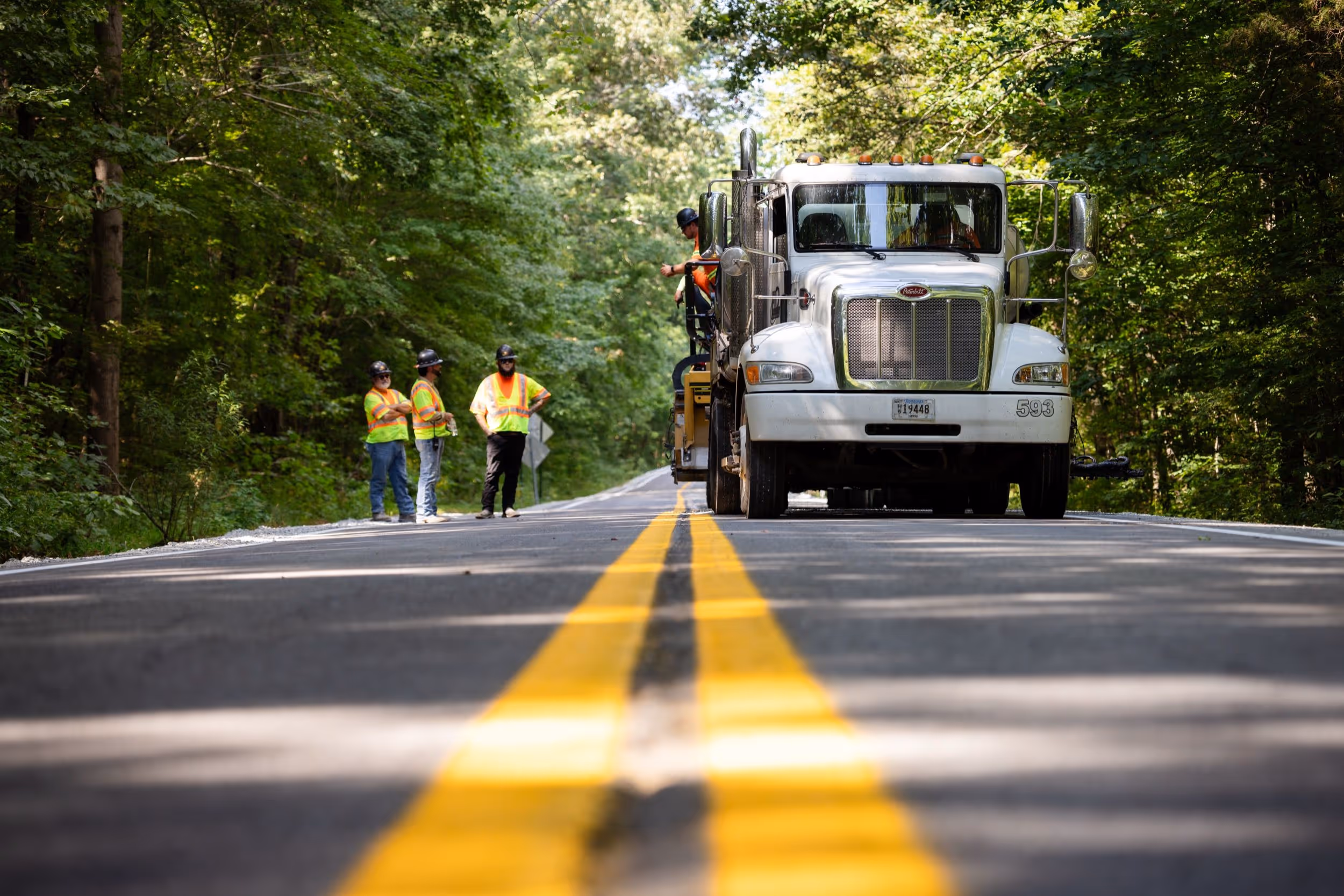 Road construction workers in high-visibility vests and helmets standing beside a large white truck on a forest-lined road with freshly painted double yellow lines.
