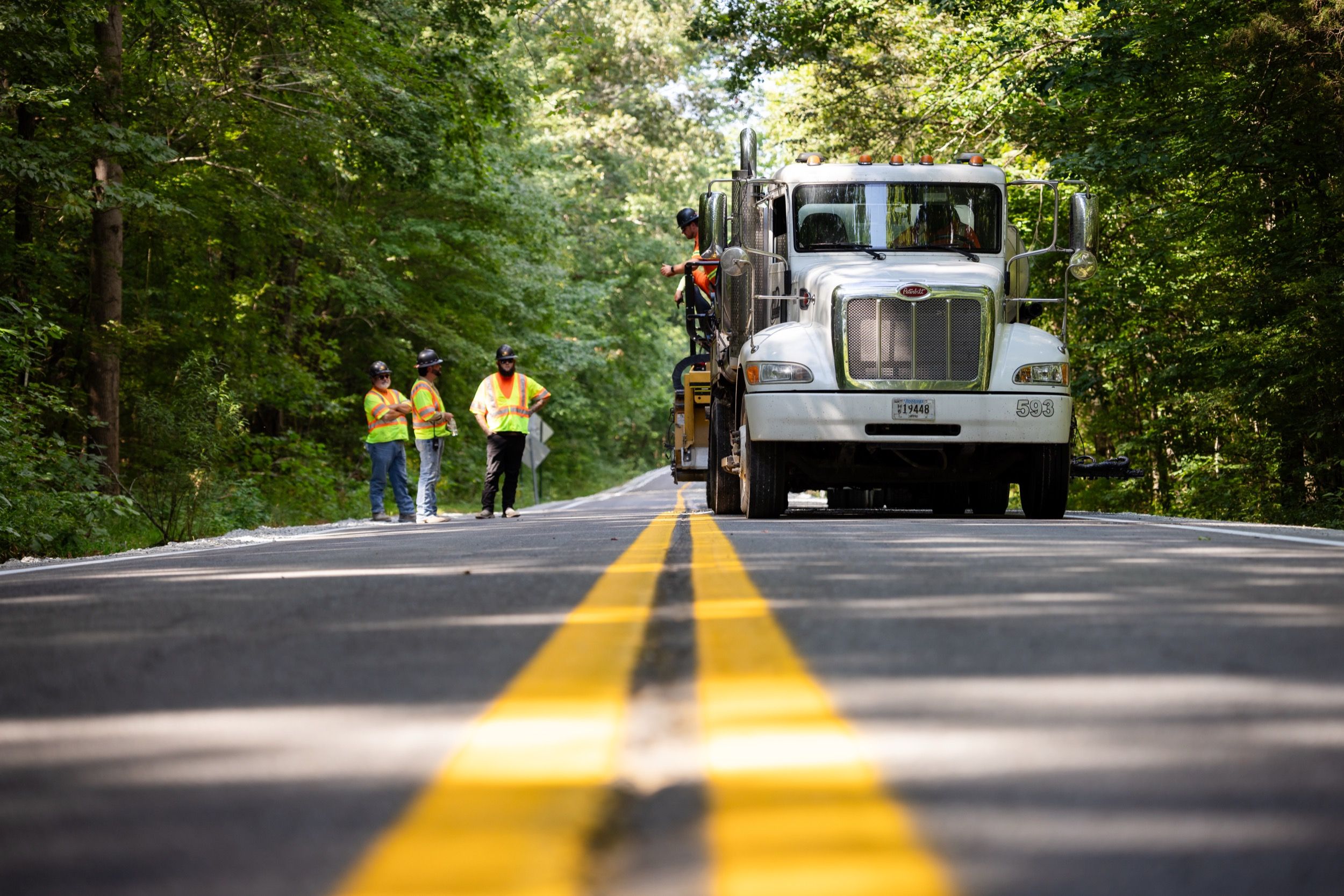 Road construction workers in high-visibility vests and helmets standing beside a large white truck on a forest-lined road with freshly painted double yellow lines.