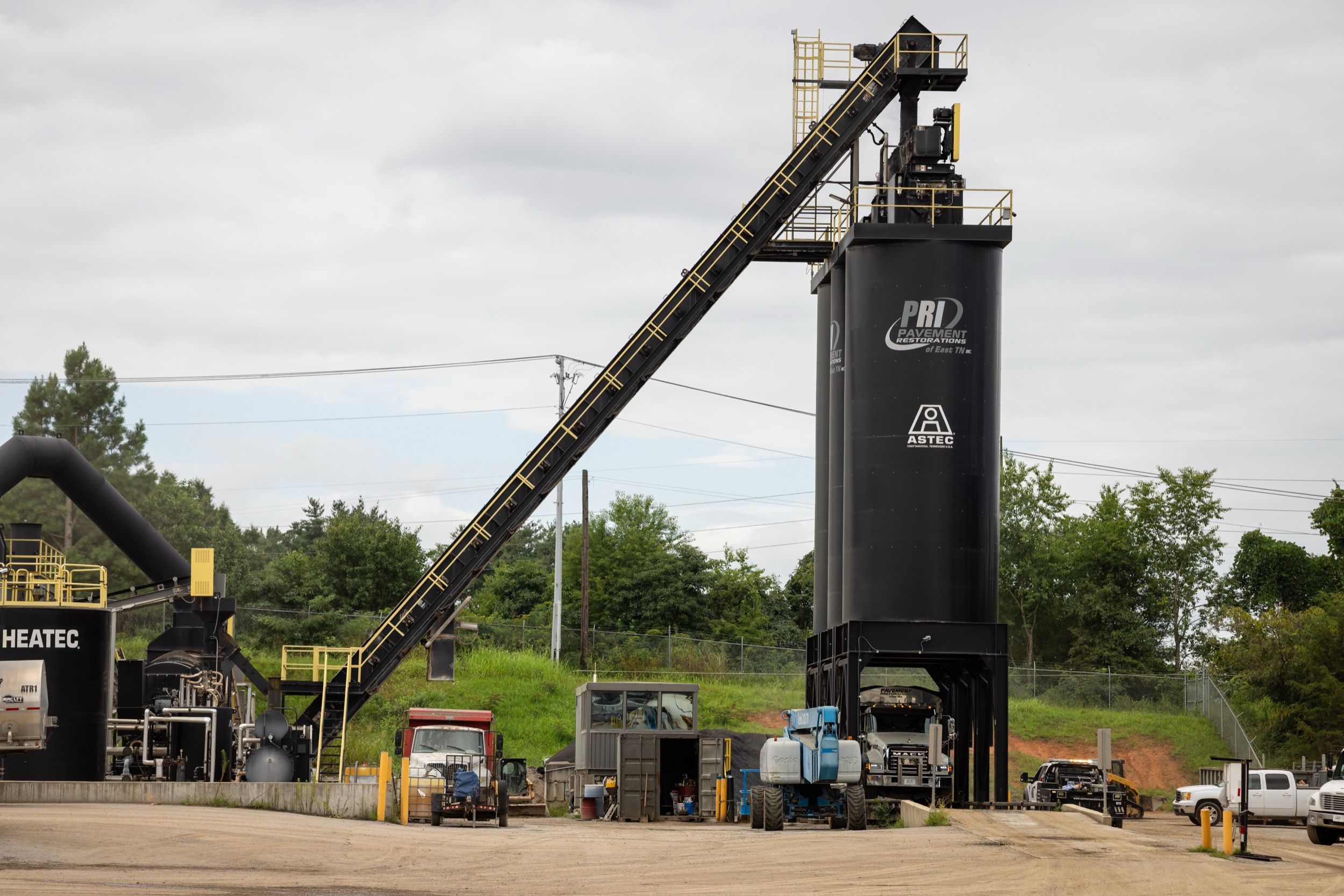 Industrial asphalt plant with tall black storage silo labeled PRI Pavement Restorations and Astec, conveyor belt, and various construction vehicles outdoors.