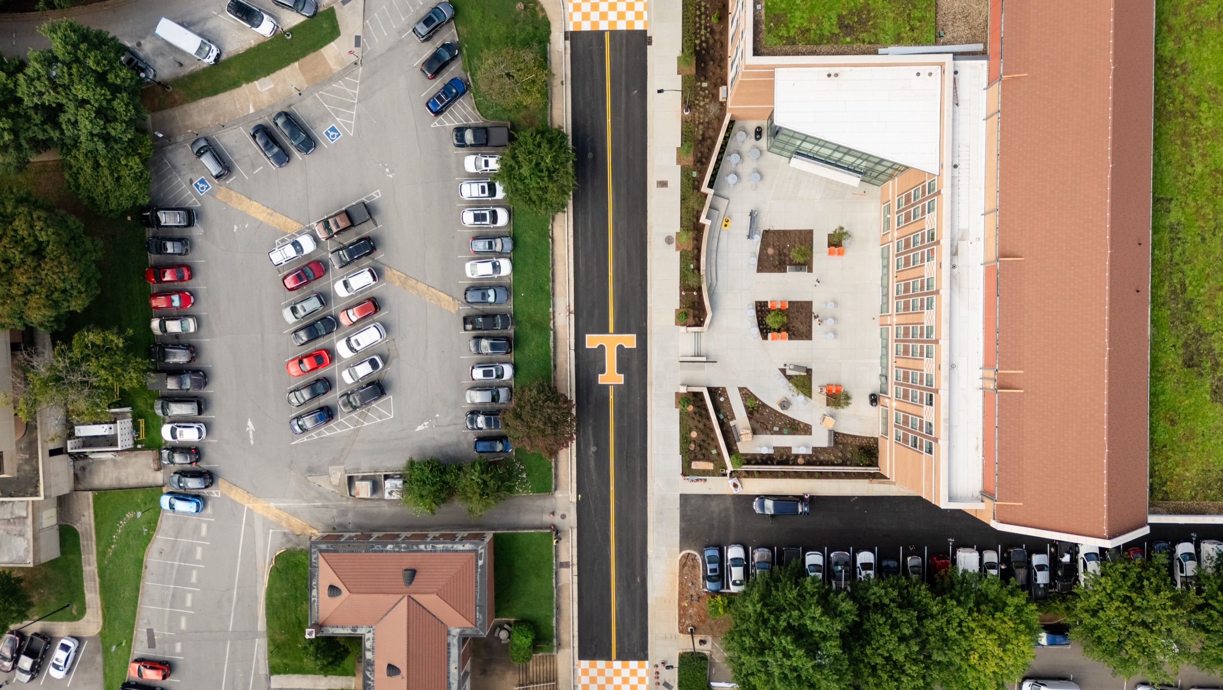 Aerial view of a parking lot with numerous parked cars next to a modern building with outdoor seating and a black road with an orange 'T' symbol in the center.