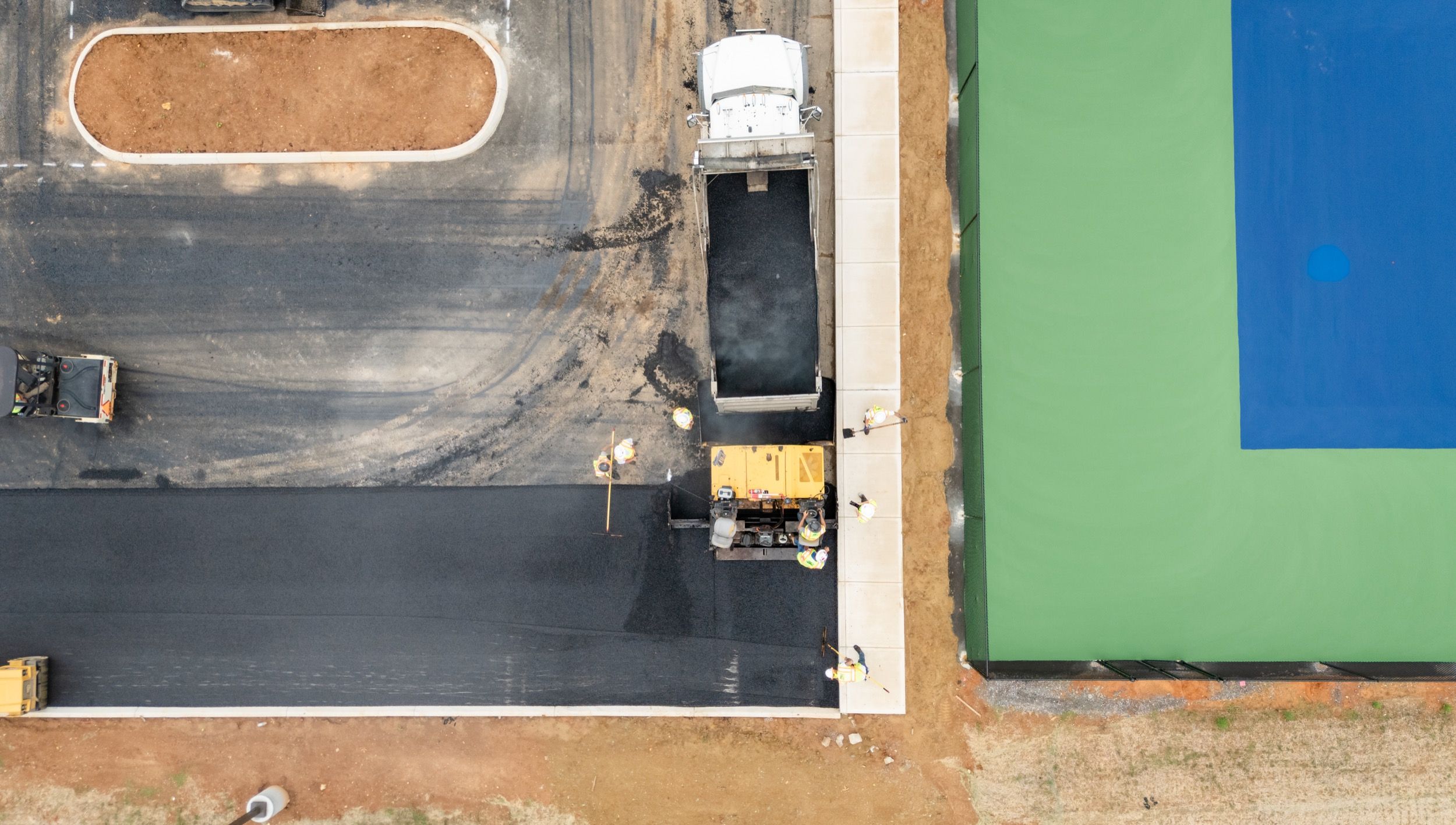 Aerial view of workers paving a black asphalt road next to a green and blue tennis court.