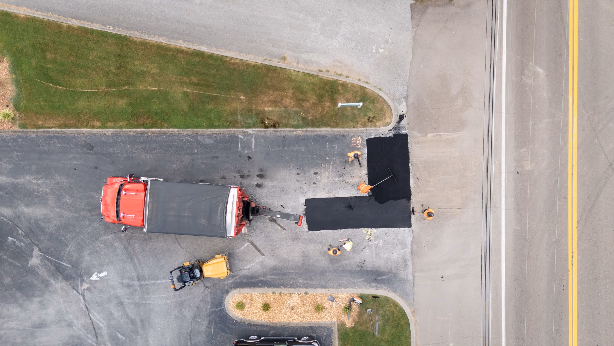 Aerial view of workers laying fresh asphalt on a driveway next to a red truck and yellow paving machine.