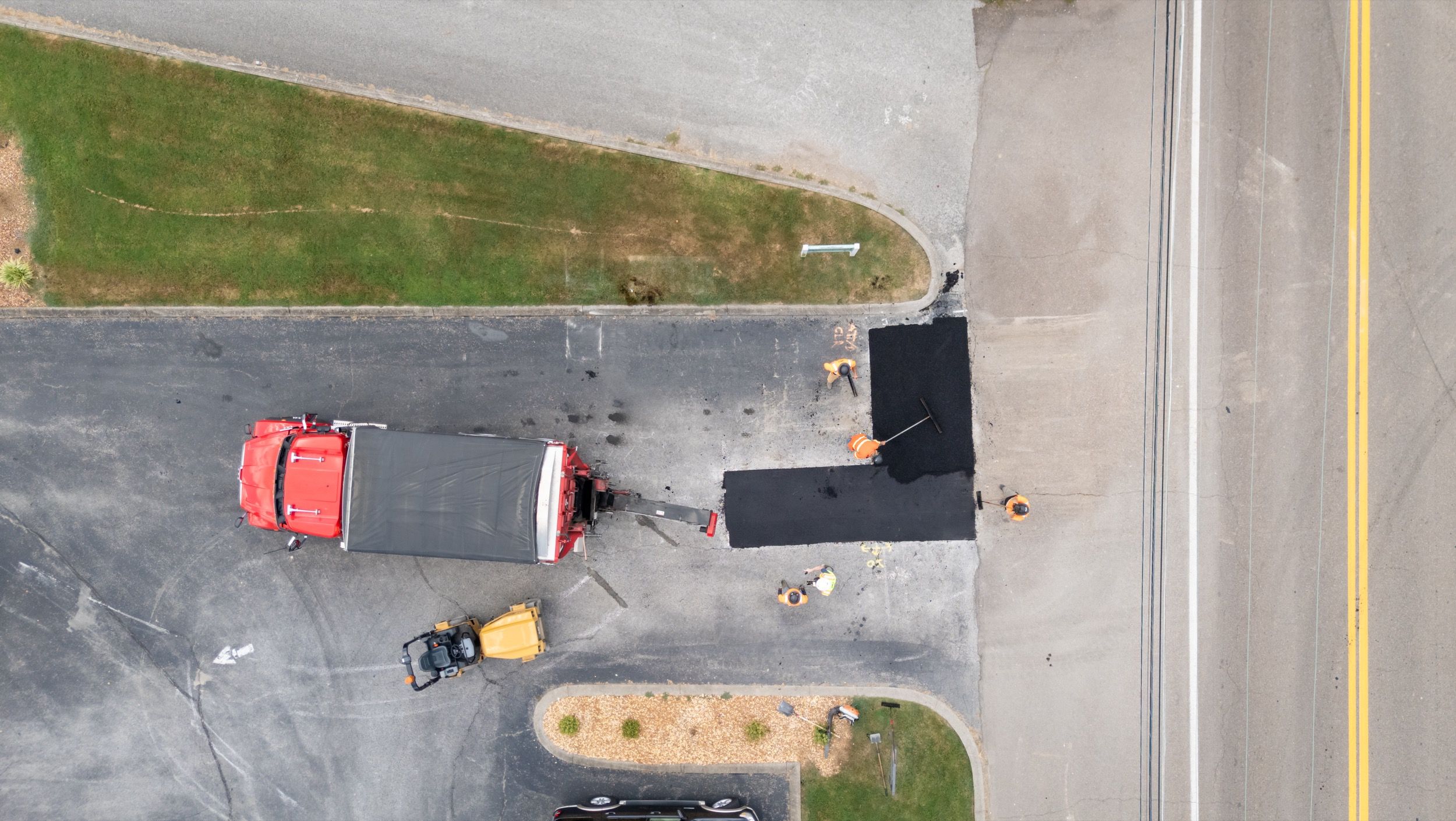 Aerial view of workers laying fresh asphalt on a driveway next to a red truck and yellow paving machine.