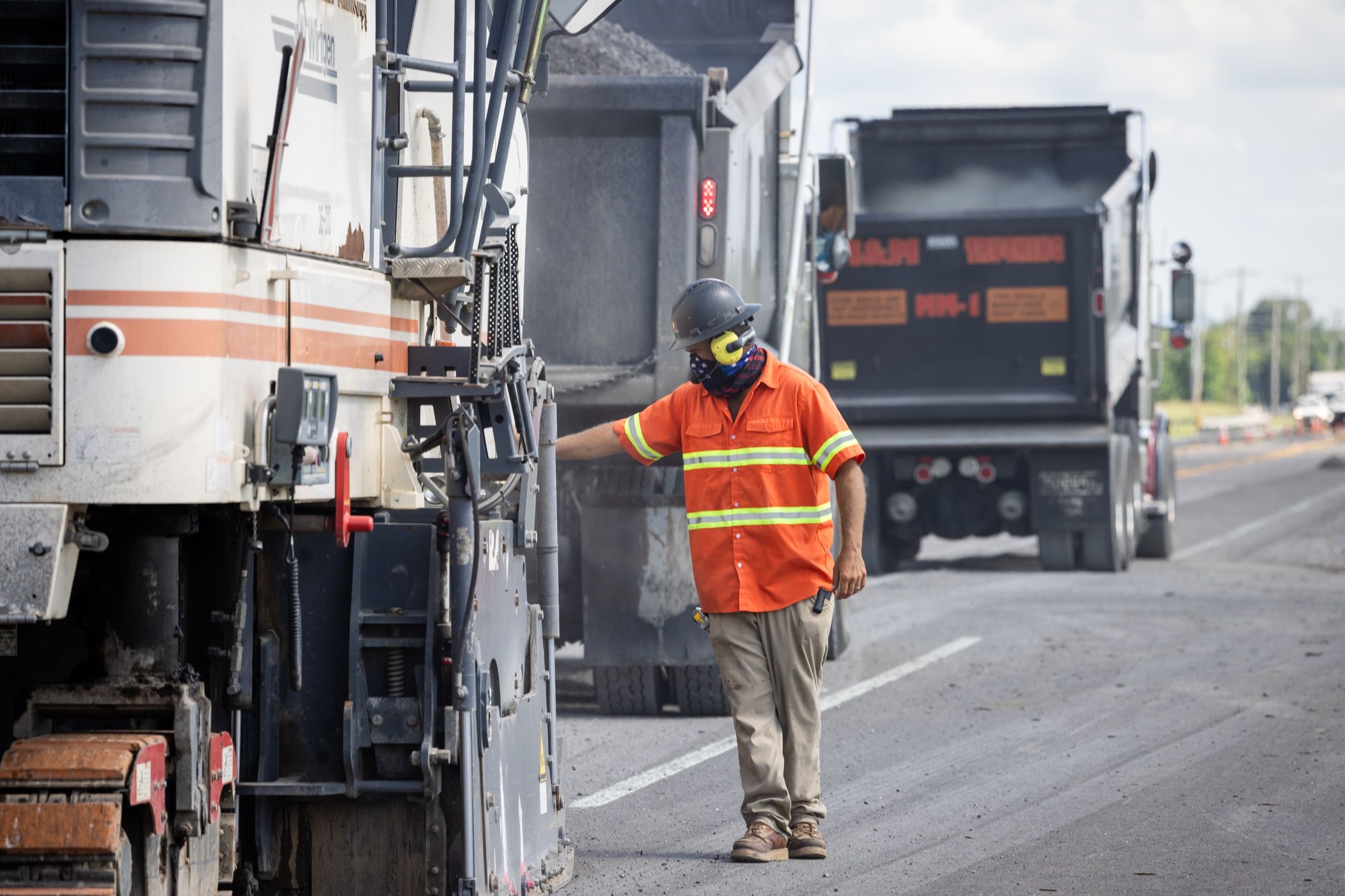 Construction worker in orange high-visibility clothing and protective gear operating road paving machinery on a highway.
