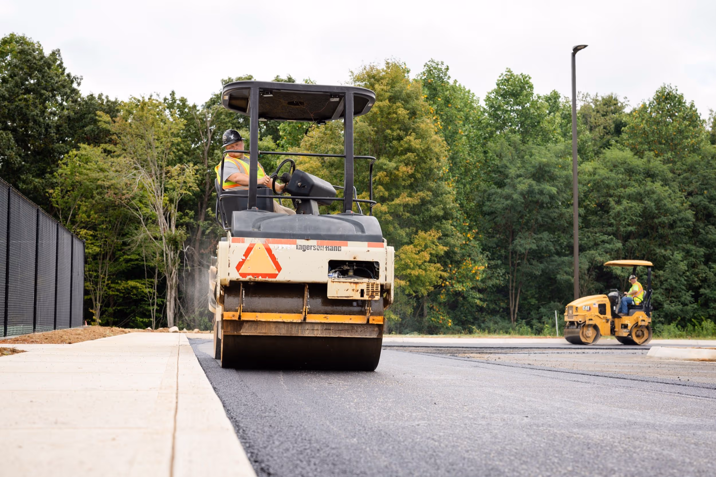 Construction workers operating road rollers to pave a new asphalt road next to a sidewalk with trees in the background.