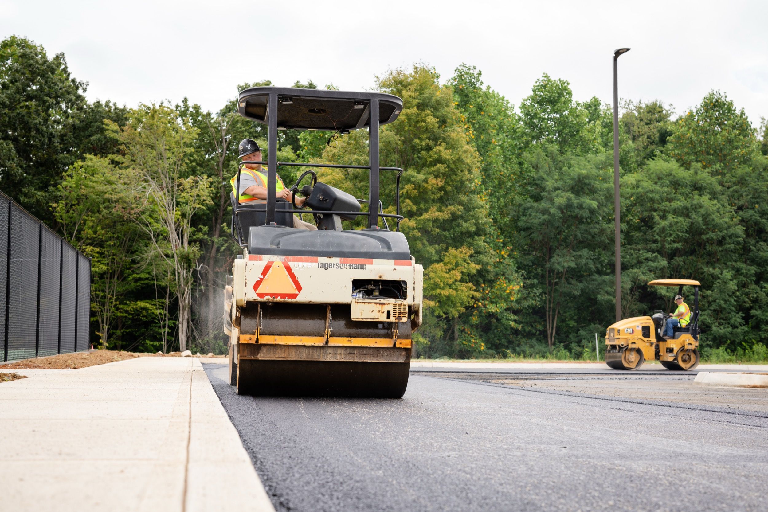 Construction workers operating road rollers to pave a new asphalt road next to a sidewalk with trees in the background.