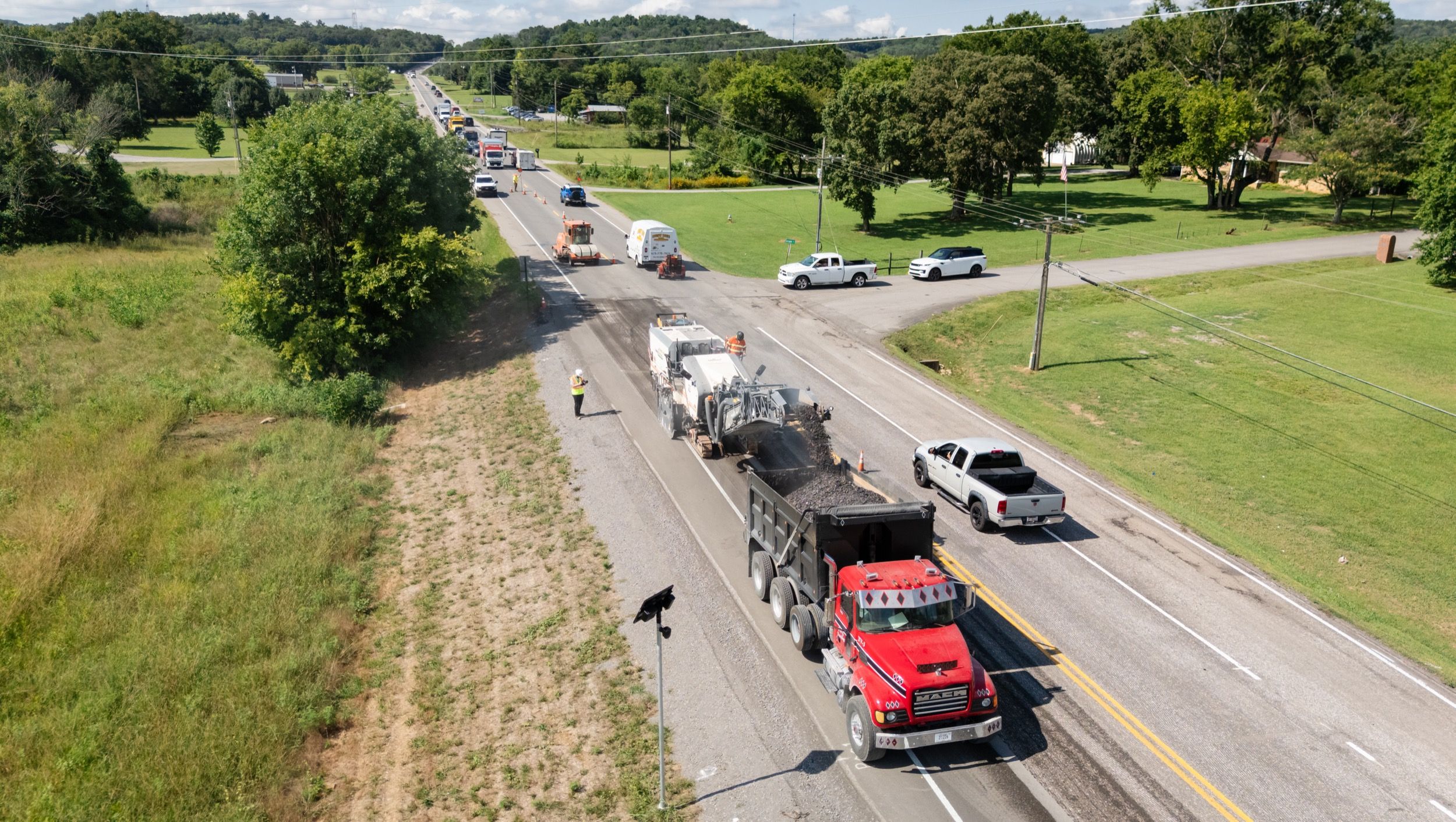 Road construction scene with a red dump truck receiving asphalt from a paving machine on a two-lane rural road.