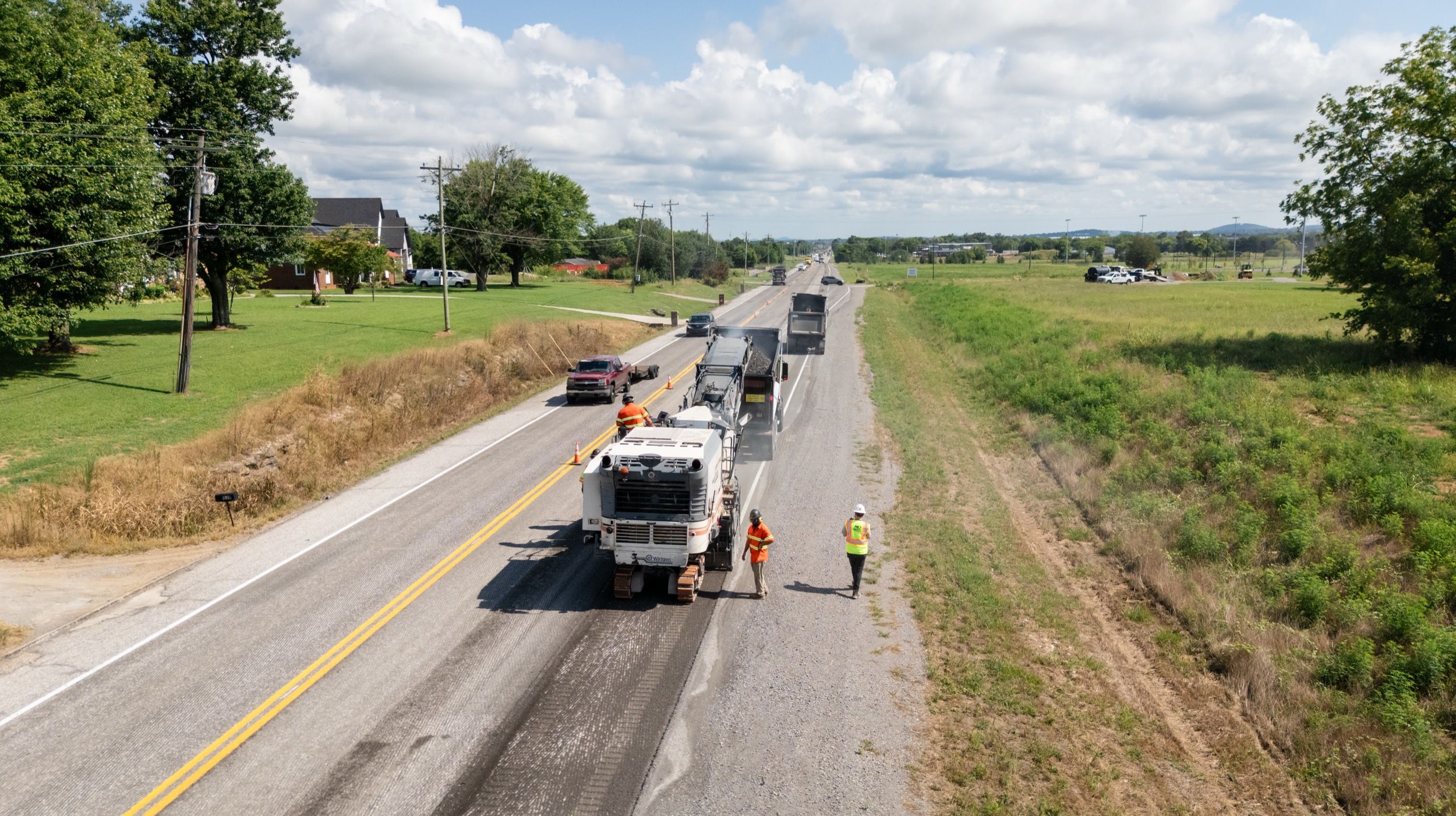 Road construction crew milling pavement on a rural highway with trucks collecting asphalt.