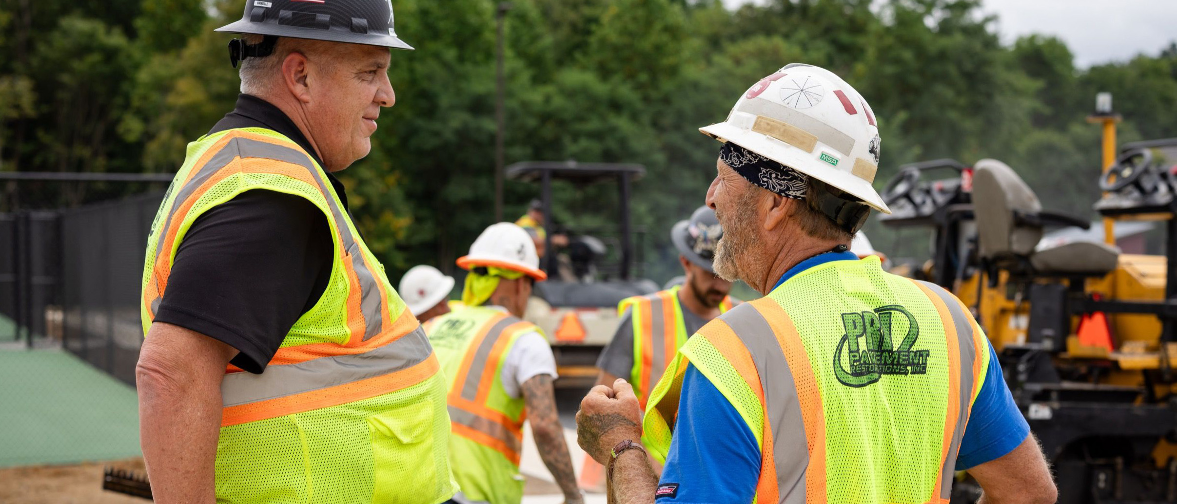Two field supervisors wearing hard hats and reflective vests discussing at a construction site with workers and equipment in the background.