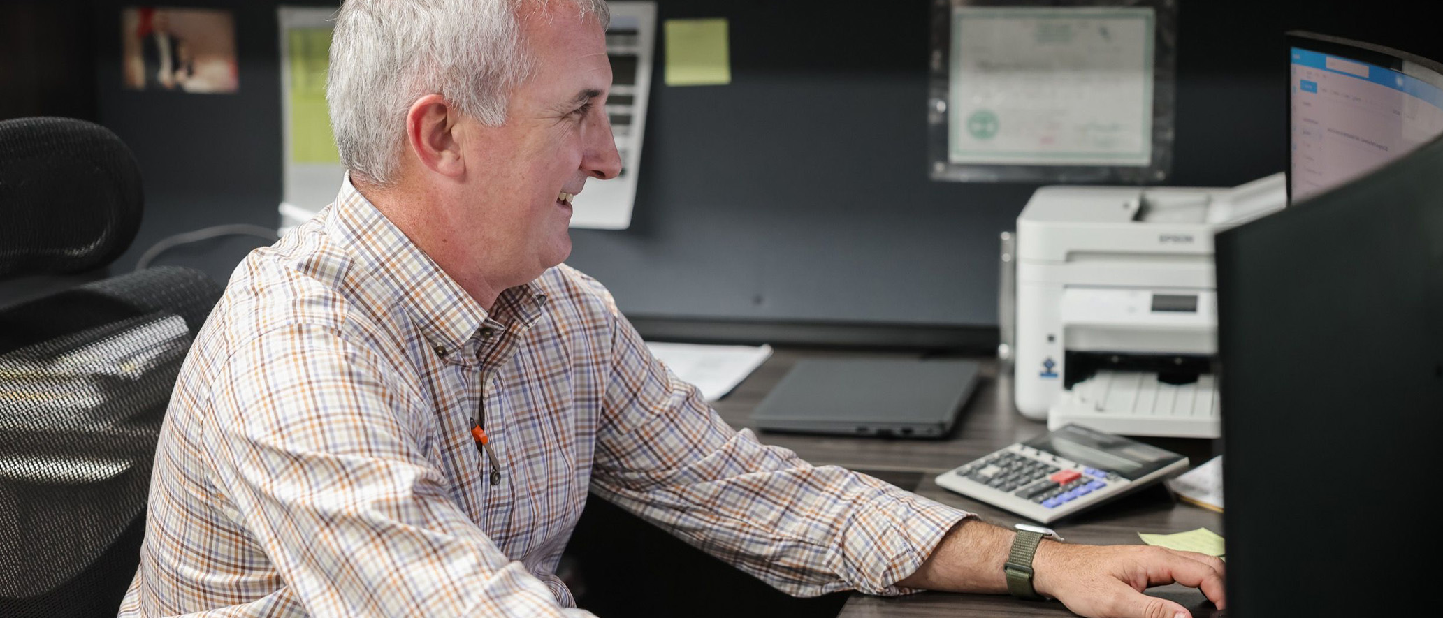 Middle-aged man in a plaid shirt smiling while working at a desk with a computer, calculator, printer, and laptop.