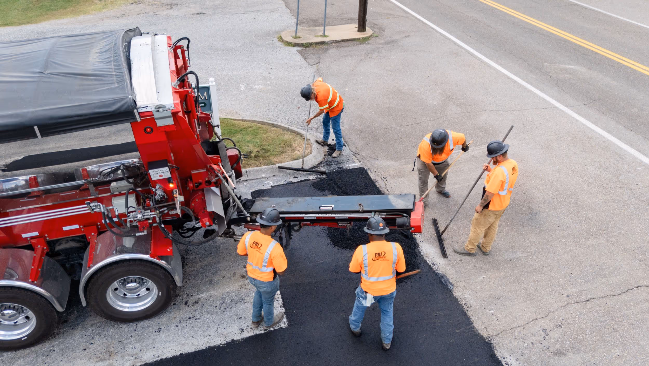 Five construction workers in orange safety vests and helmets paving a road with hot asphalt from a red paving truck.
