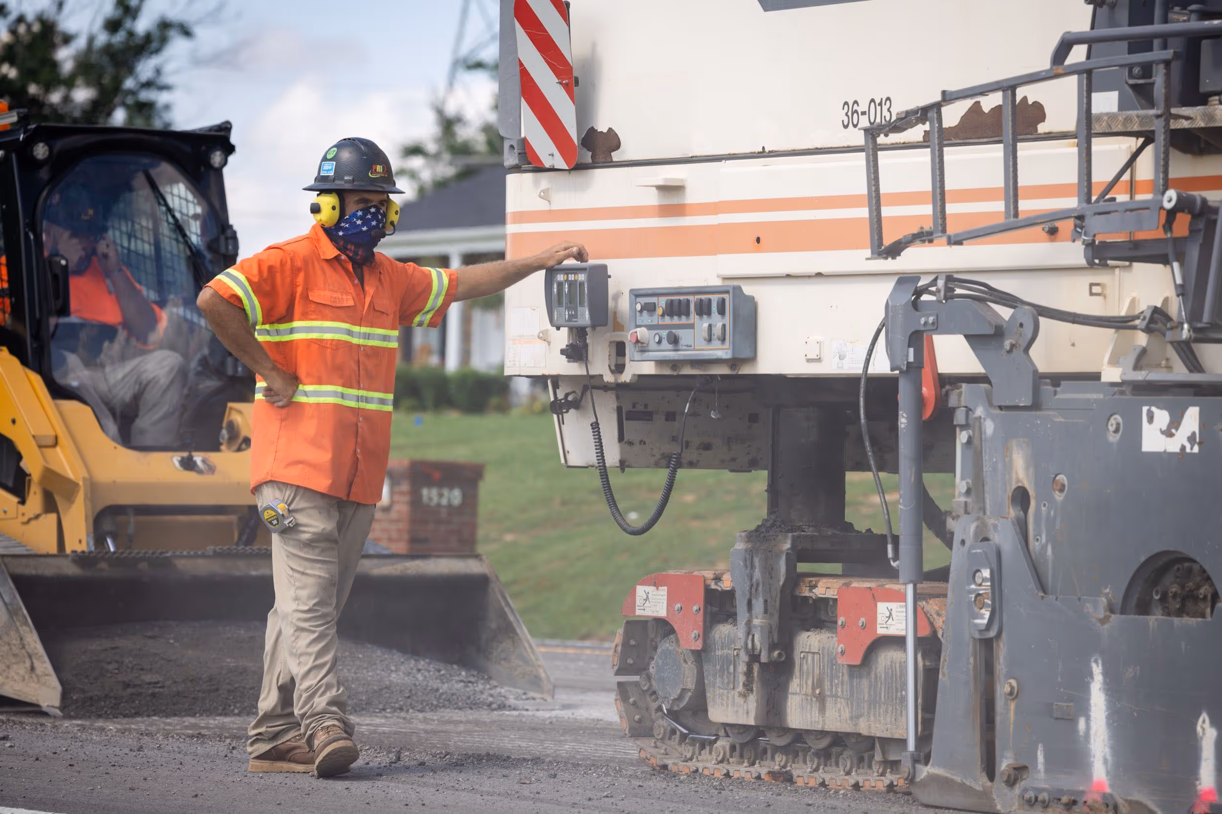 Construction worker in orange safety shirt and helmet with earmuffs standing next to heavy machinery on a roadwork site.