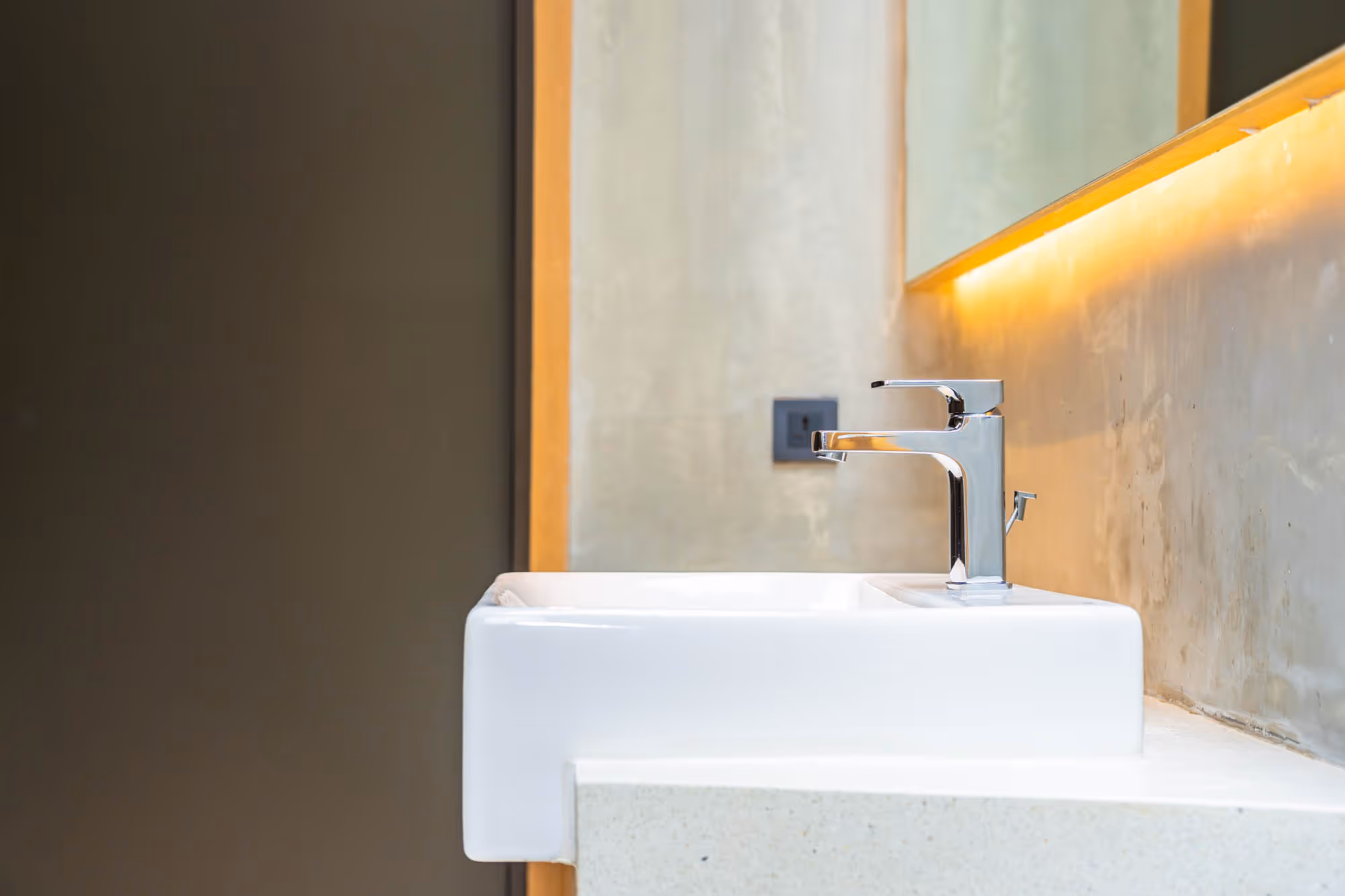 A modern rectangular sink with a chrome faucet is installed against a beige wall with a mirror.