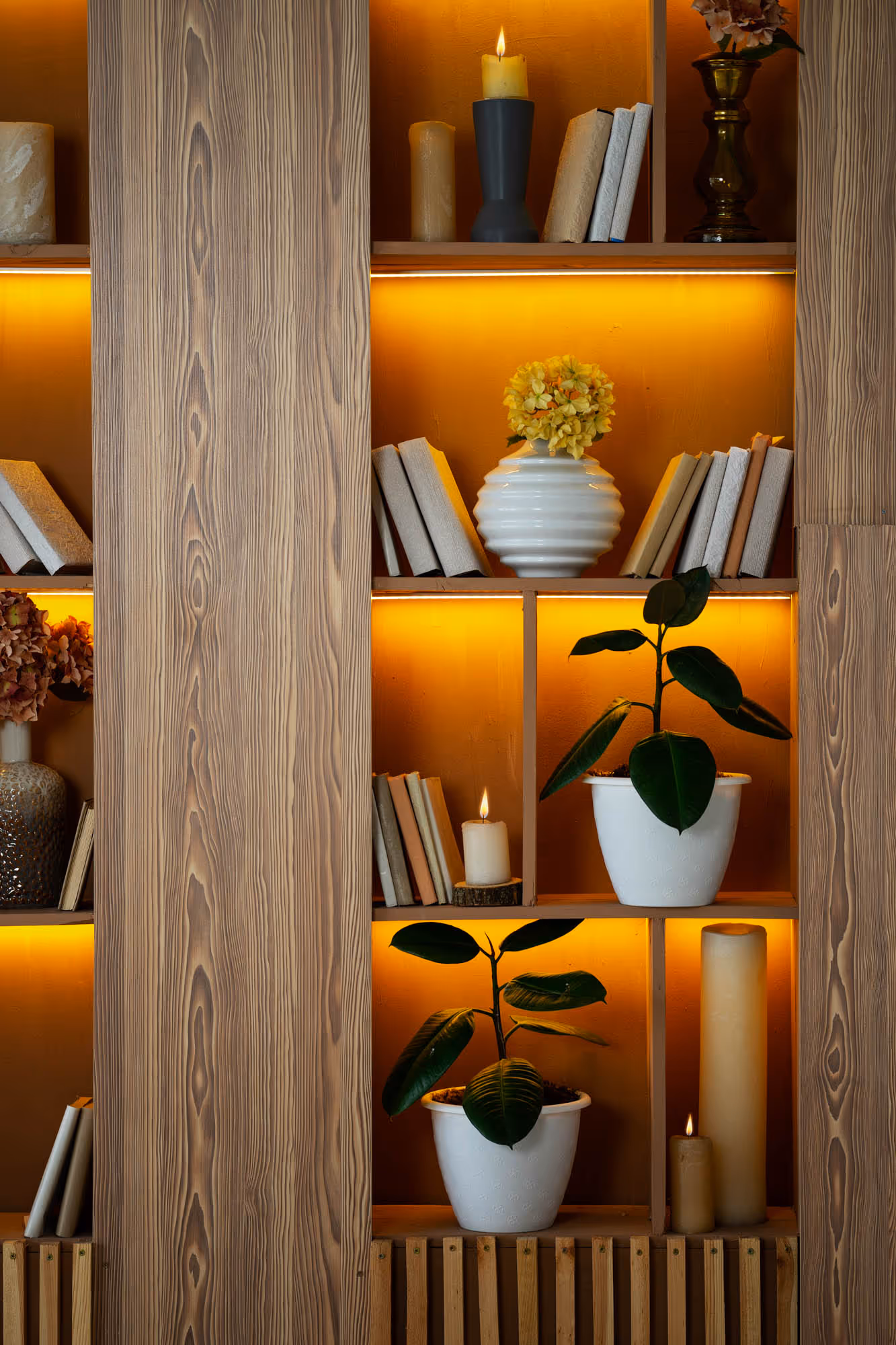 Wooden shelves with books, candles, and potted plants. A vase holds yellow flowers on the top shelf.