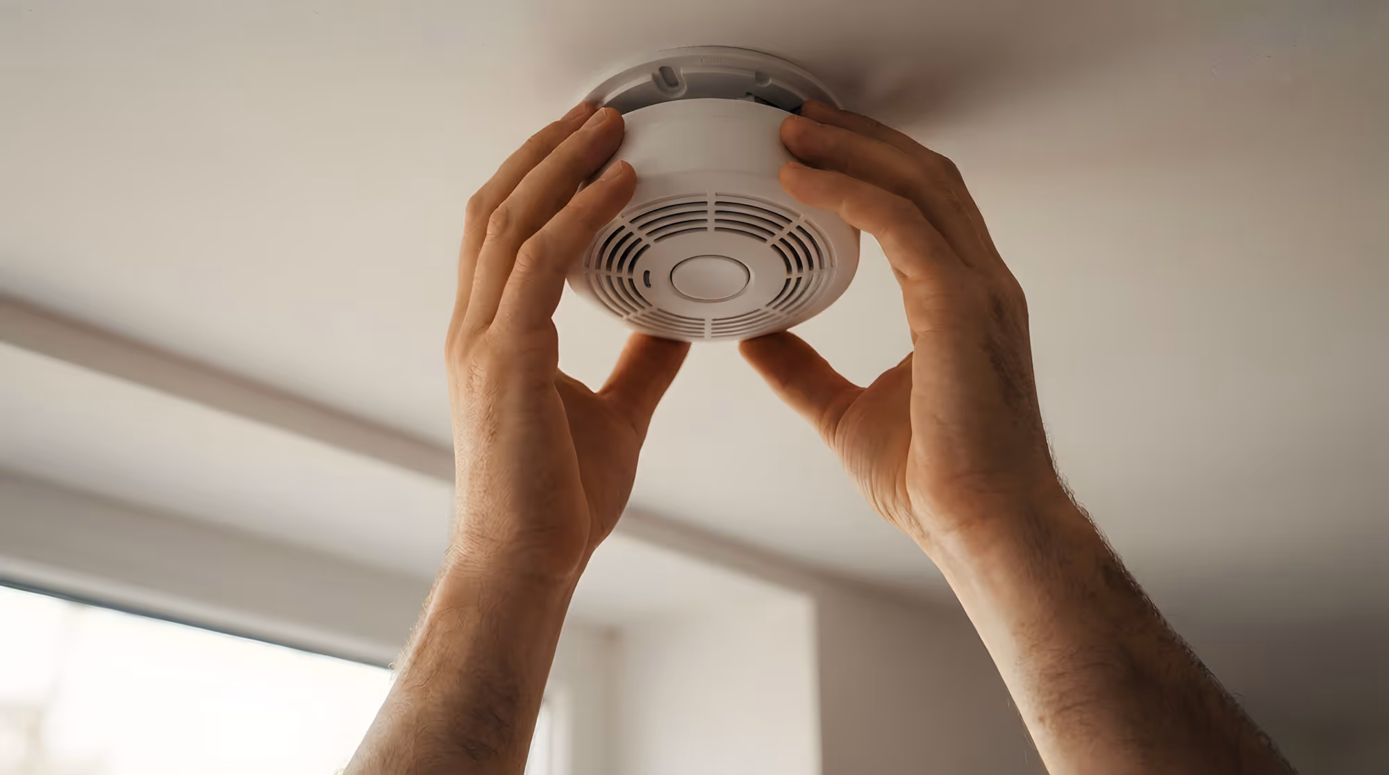 Hands installing a smoke detector on a ceiling.