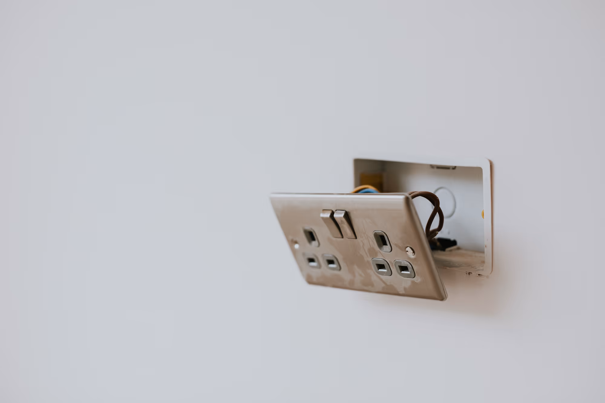 A partially detached electrical outlet with visible wires hangs from a white wall.