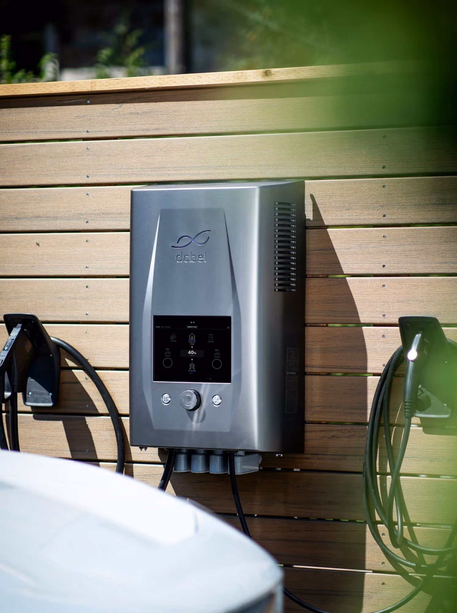 A wall-mounted gray electric vehicle charging station with cables hangs on a wooden fence.