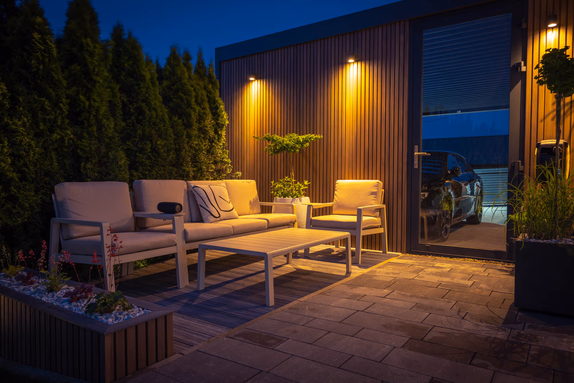 Outdoor patio with beige seating, a table, and warm lighting on a wooden deck at night.