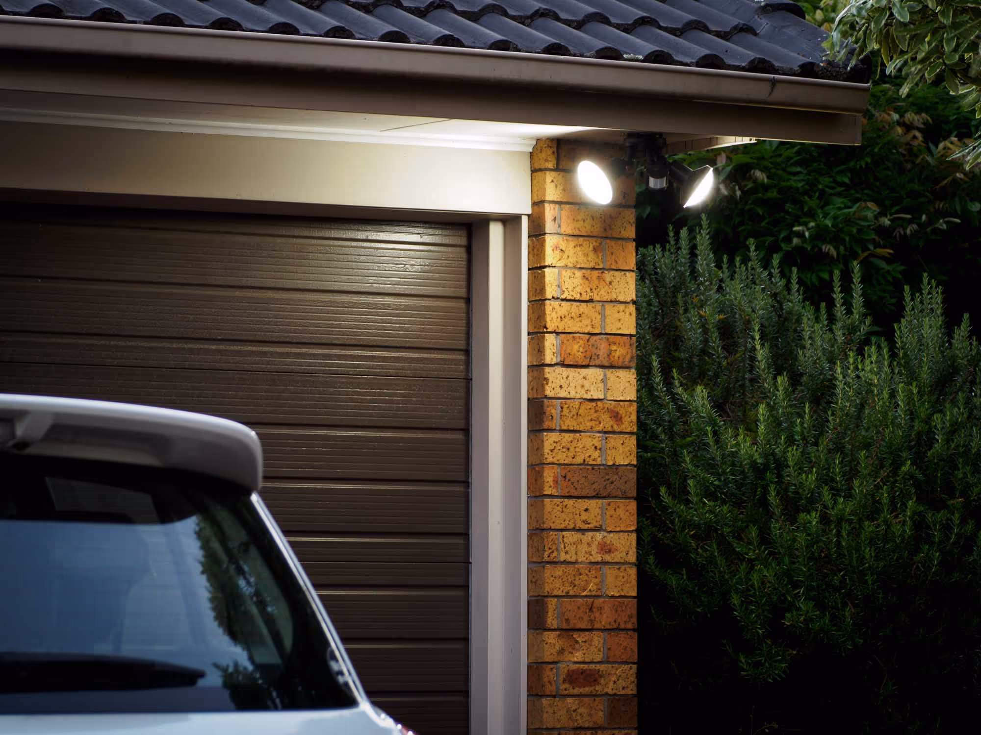 A parked car is in front of a garage with two security lights on a brick wall.