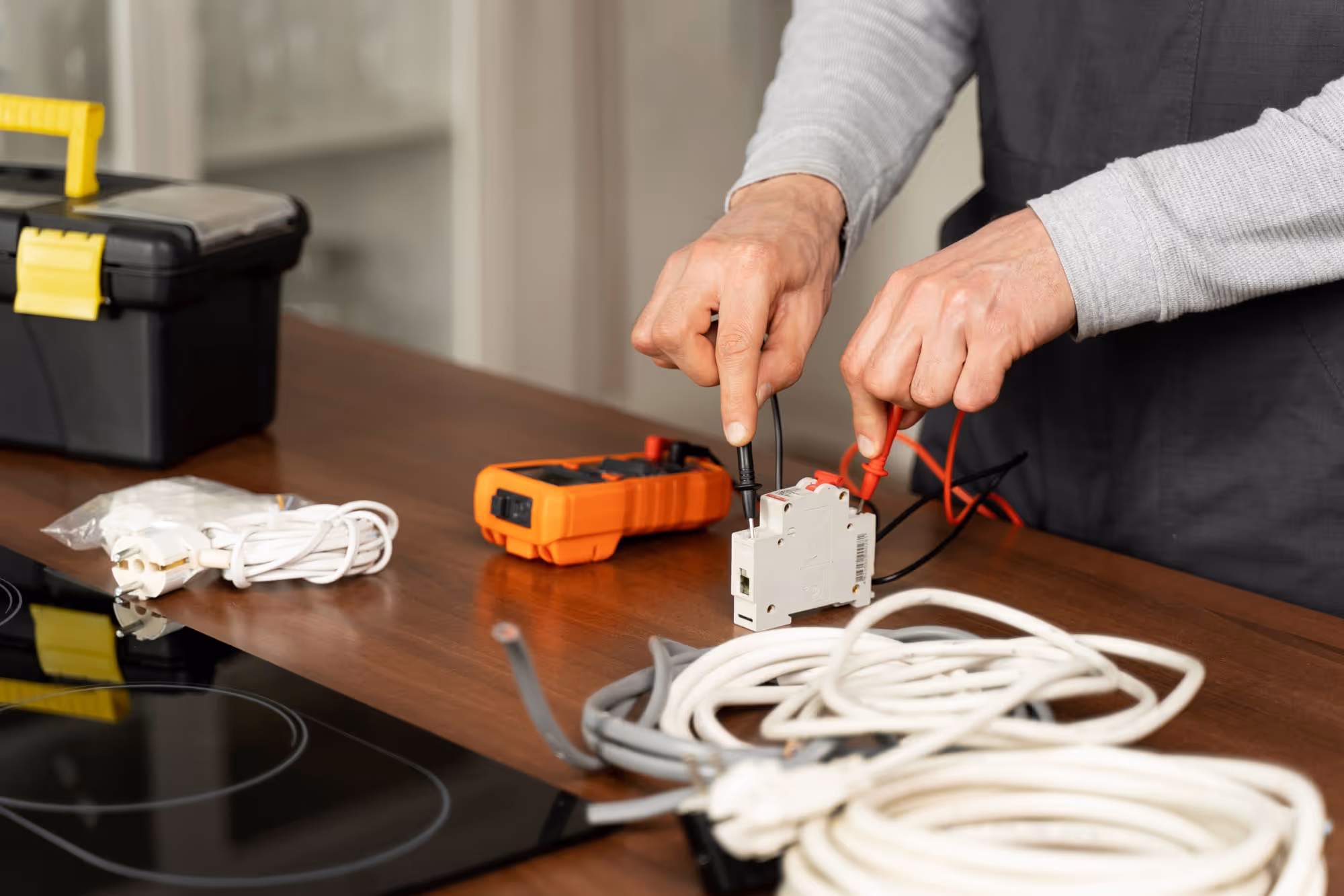 Person testing an electrical device with a multimeter on a wooden table, surrounded by wires and tools.