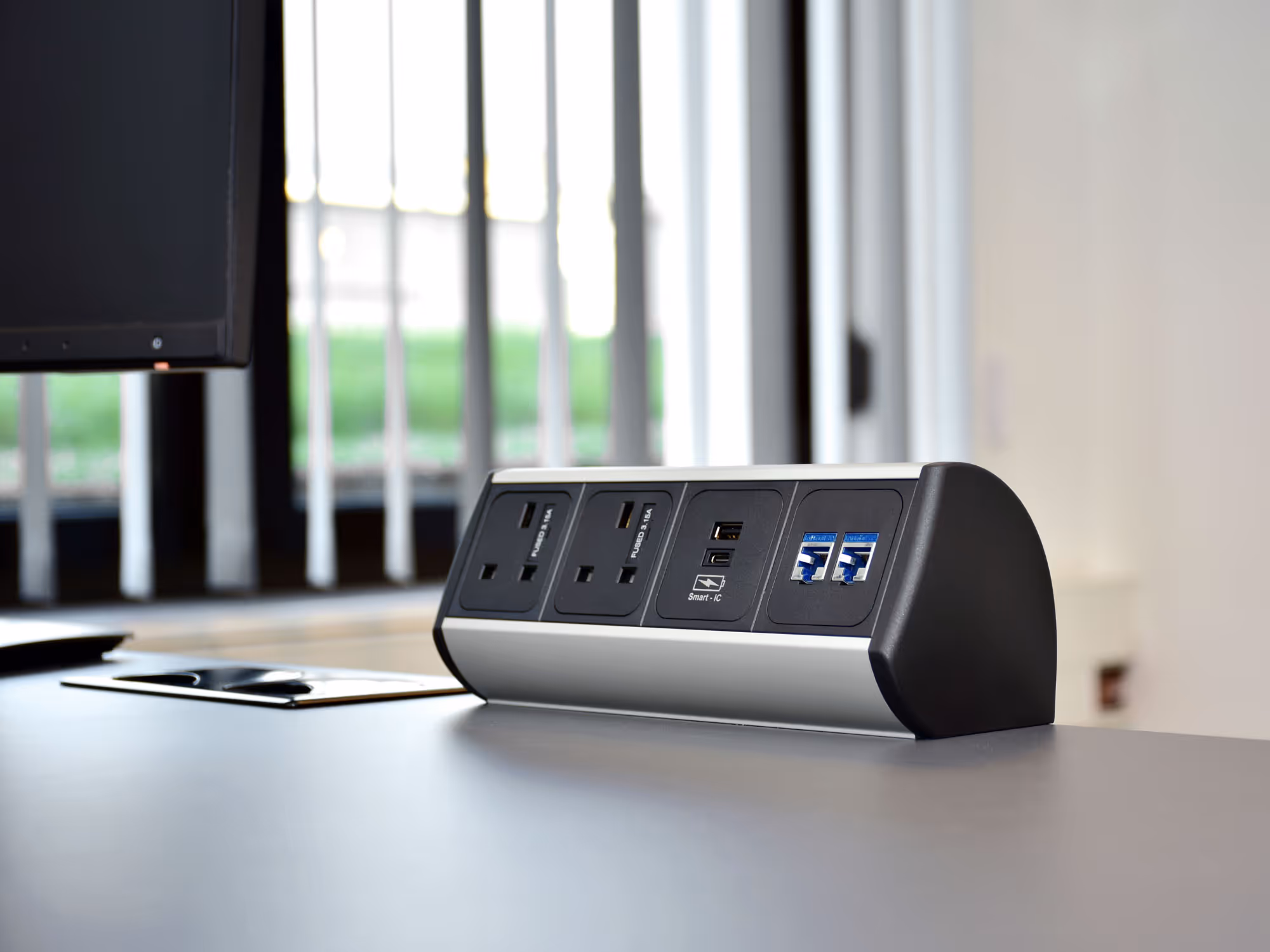 A power strip with electrical outlets and USB ports is placed on a desk near a window.
