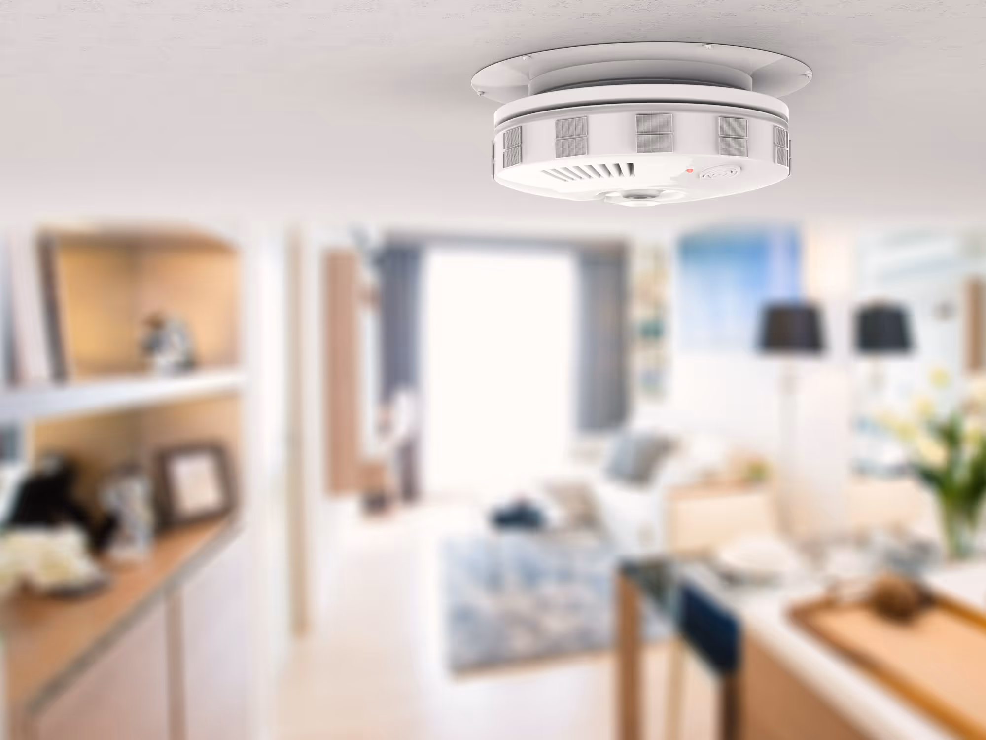 A smoke detector mounted on the ceiling in a modern, blurred living room setting.