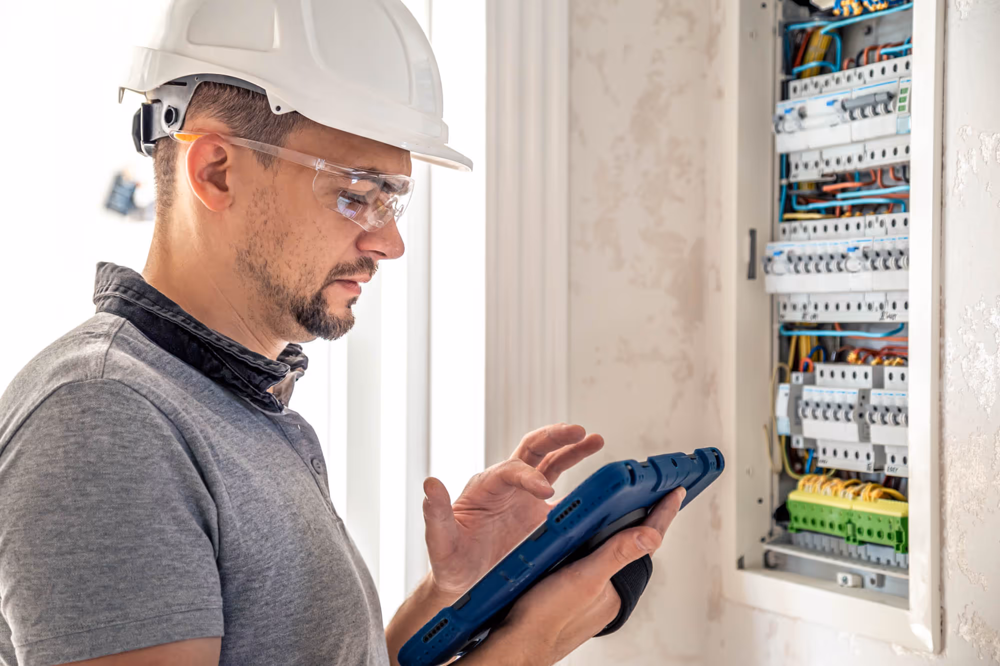Electrician wearing safety gear uses a tablet to inspect a circuit breaker panel.