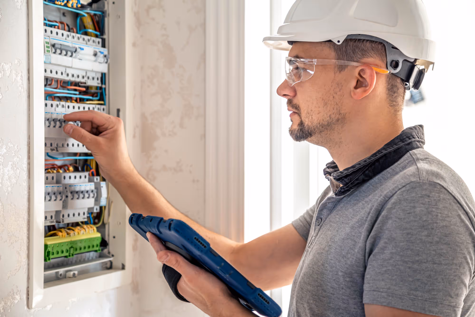 A technician with a helmet checks wiring in an electrical panel while holding a tablet.