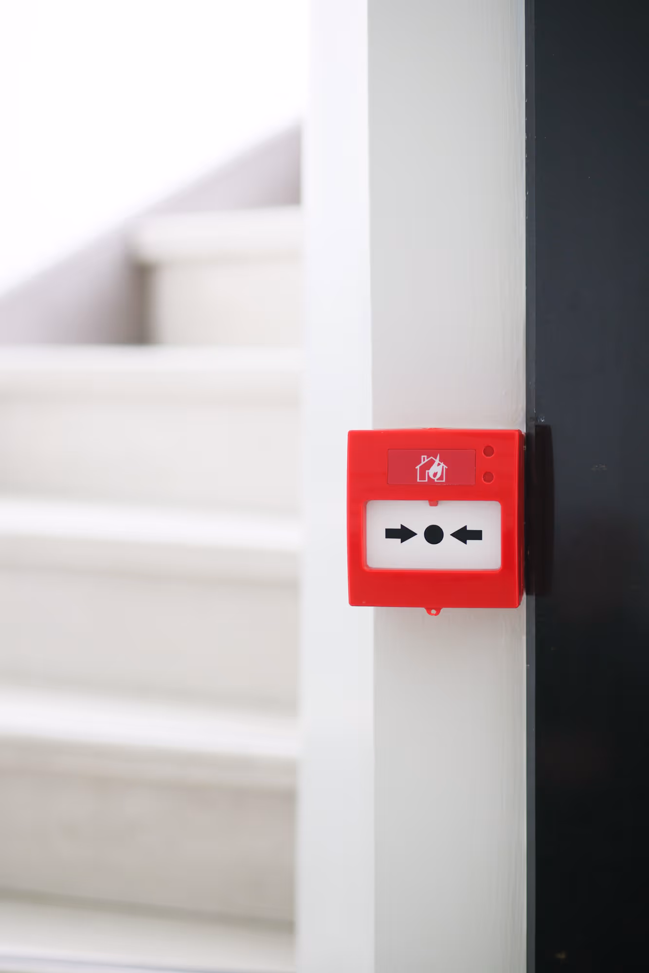 A red fire alarm is mounted on a white wall near a stairway.
