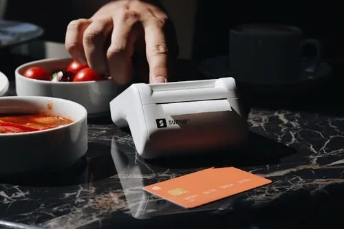 This photo showcases the SumUp Solo Printer card reader on a restaurant table, surrounded by food and coffee. It's perfect for small businesses and entrepreneurs, offering seamless payment processing in cafes and restaurants. Its sleek design and portability make it perfect for handling on-the-go transactions.