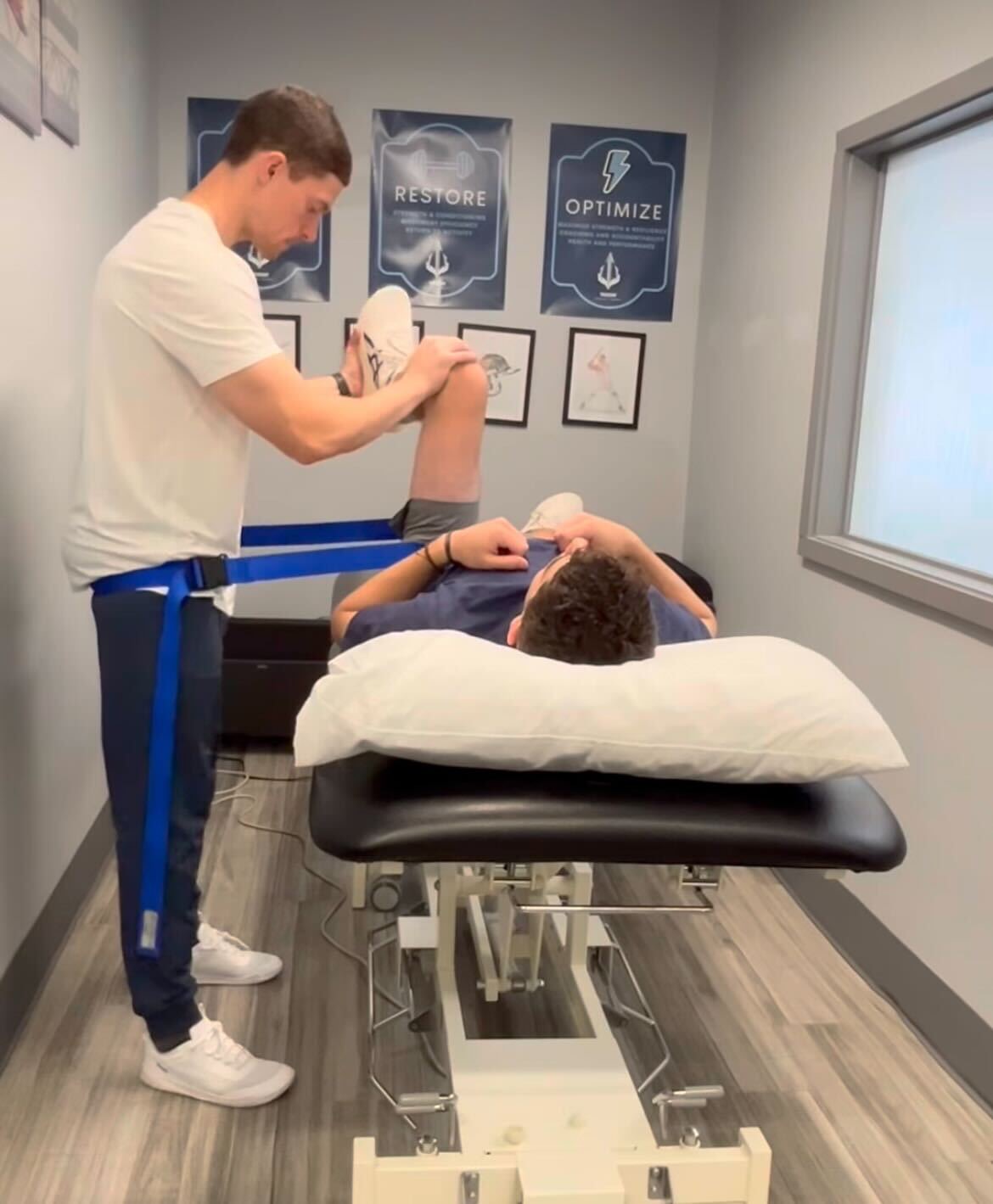 A therapist wearing a resistance band assists a person lying on a treatment table with a knee stretch exercise in a clinical room.