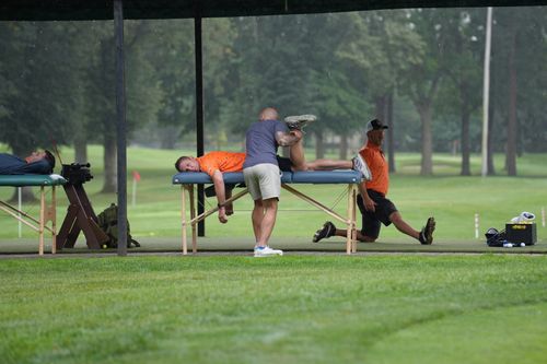 A man in a grey shirt stretches the leg of a man lying face down on a massage table outdoors on a golf course, while another man stretches nearby.