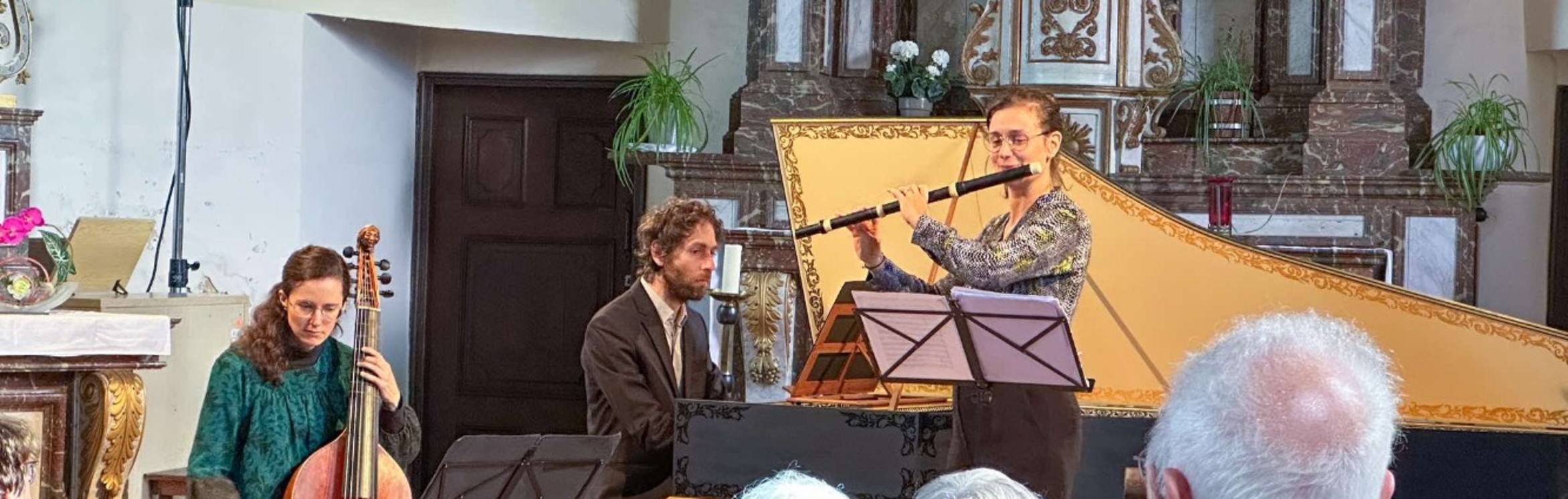 Orchestra and choir performing on stage with conductor in spotlight, surrounded by string instruments and harpsichord.