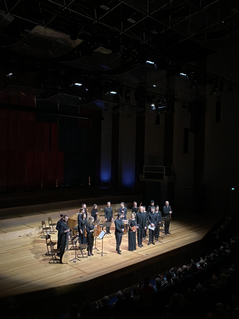 Orchestra and choir performing on stage with conductor in spotlight, surrounded by string instruments and harpsichord.