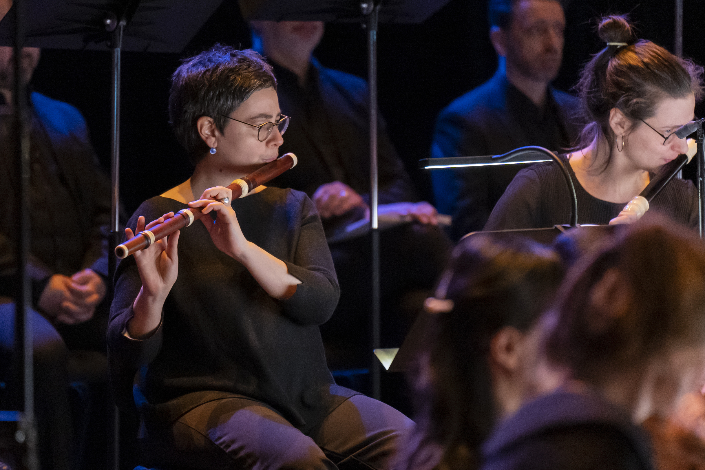 Orchestra and choir performing on stage with conductor in spotlight, surrounded by string instruments and harpsichord.