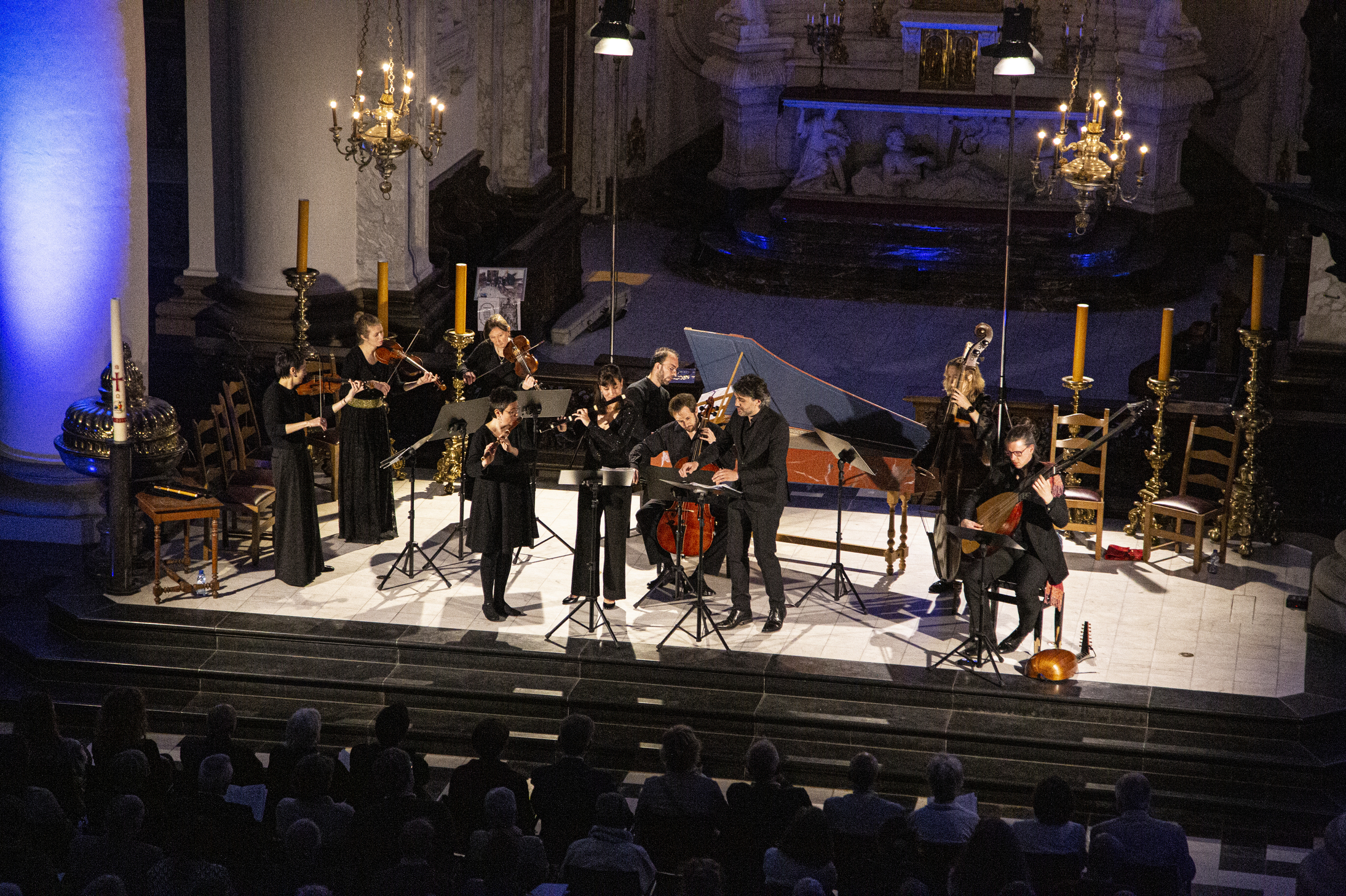 Orchestra and choir performing on stage with conductor in spotlight, surrounded by string instruments and harpsichord.
