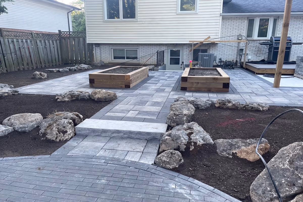 Backyard patio with gray stone pavers, two wooden raised garden beds, and large decorative rocks surrounding soil beds.