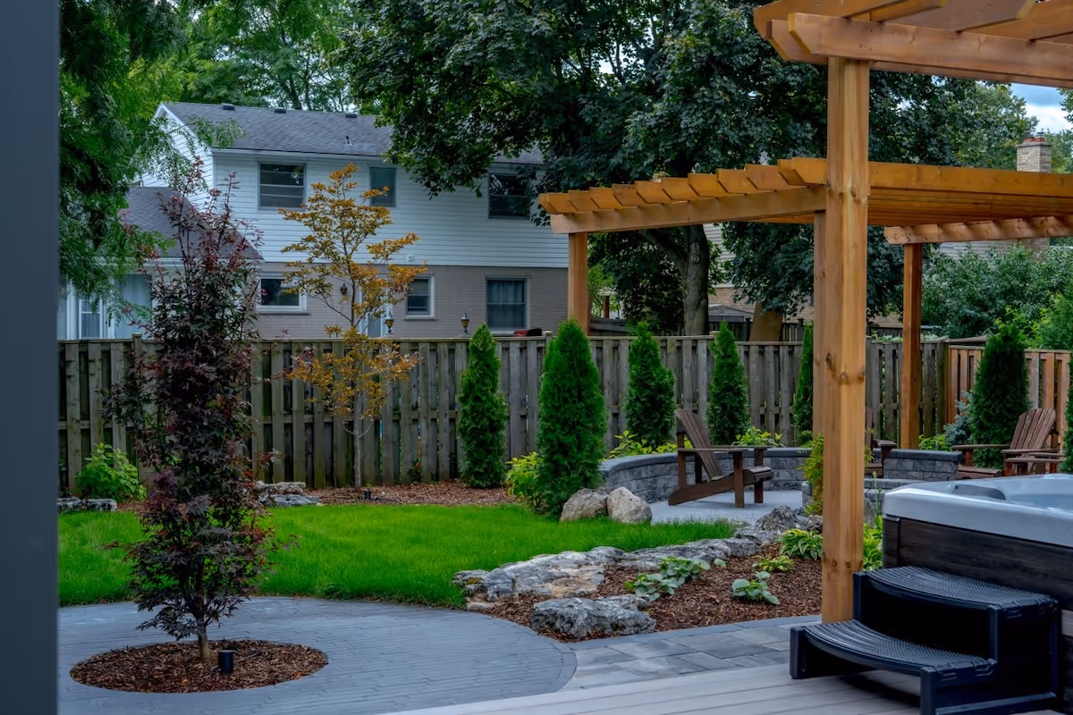 Backyard with wooden pergola, hot tub with steps, green lawn, stone fire pit surrounded by Adirondack chairs, and a wooden fence with trees.