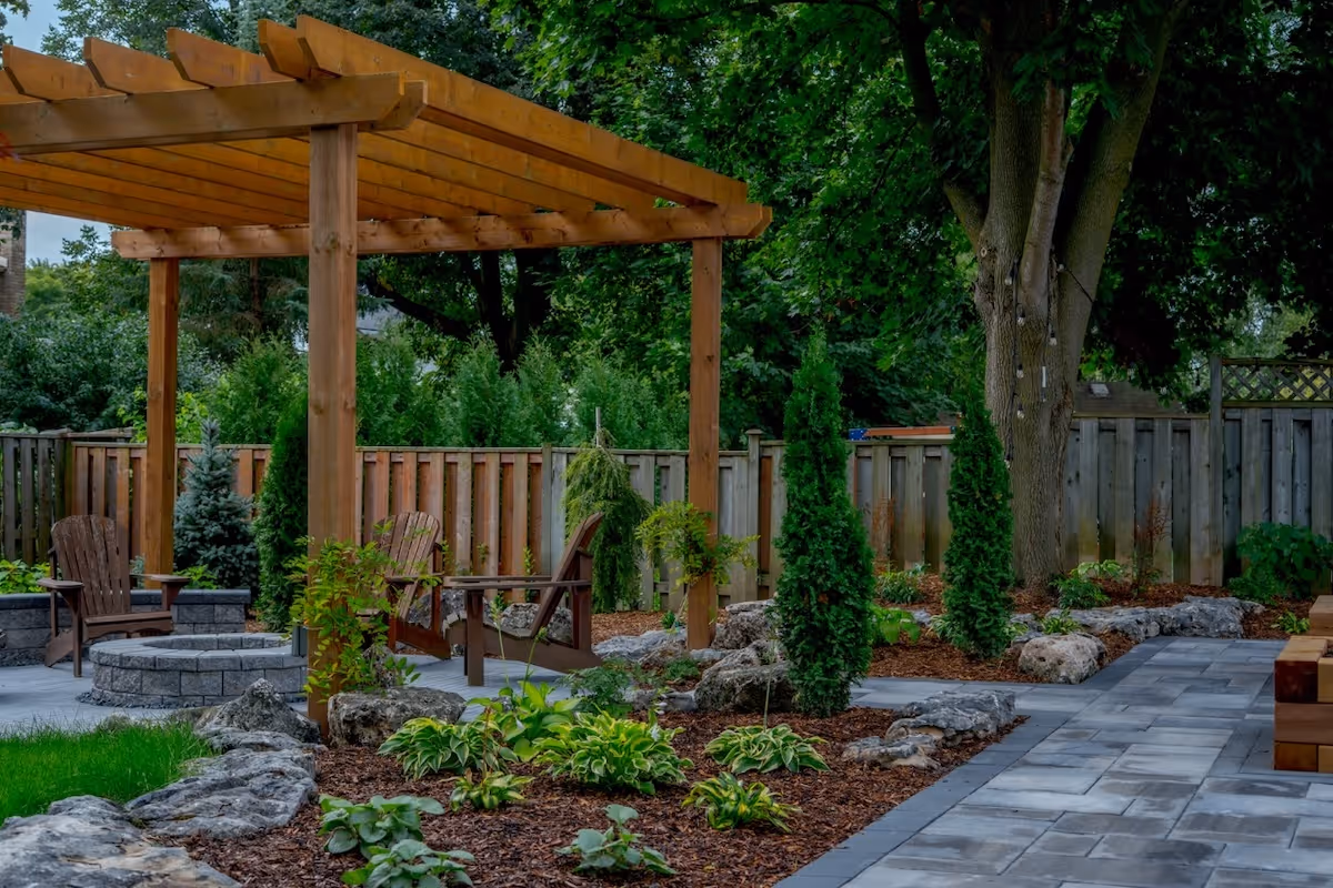 Backyard patio with wooden pergola, Adirondack chairs around a stone fire pit, landscaped garden beds, and a paved stone walkway.
