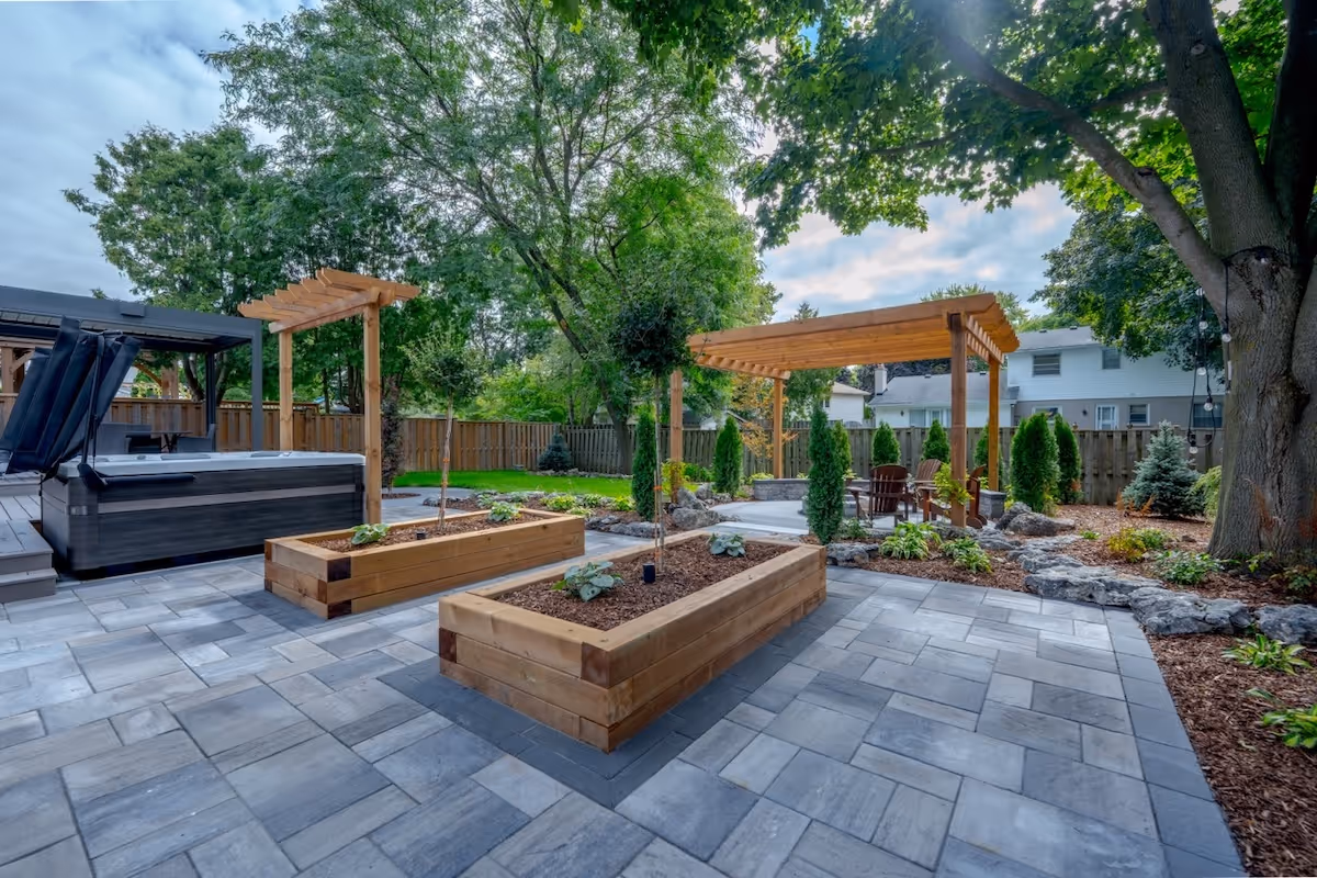 Backyard patio with large gray pavers, two wooden raised garden beds, a hot tub, a wooden pergola seating area, and lush green trees.