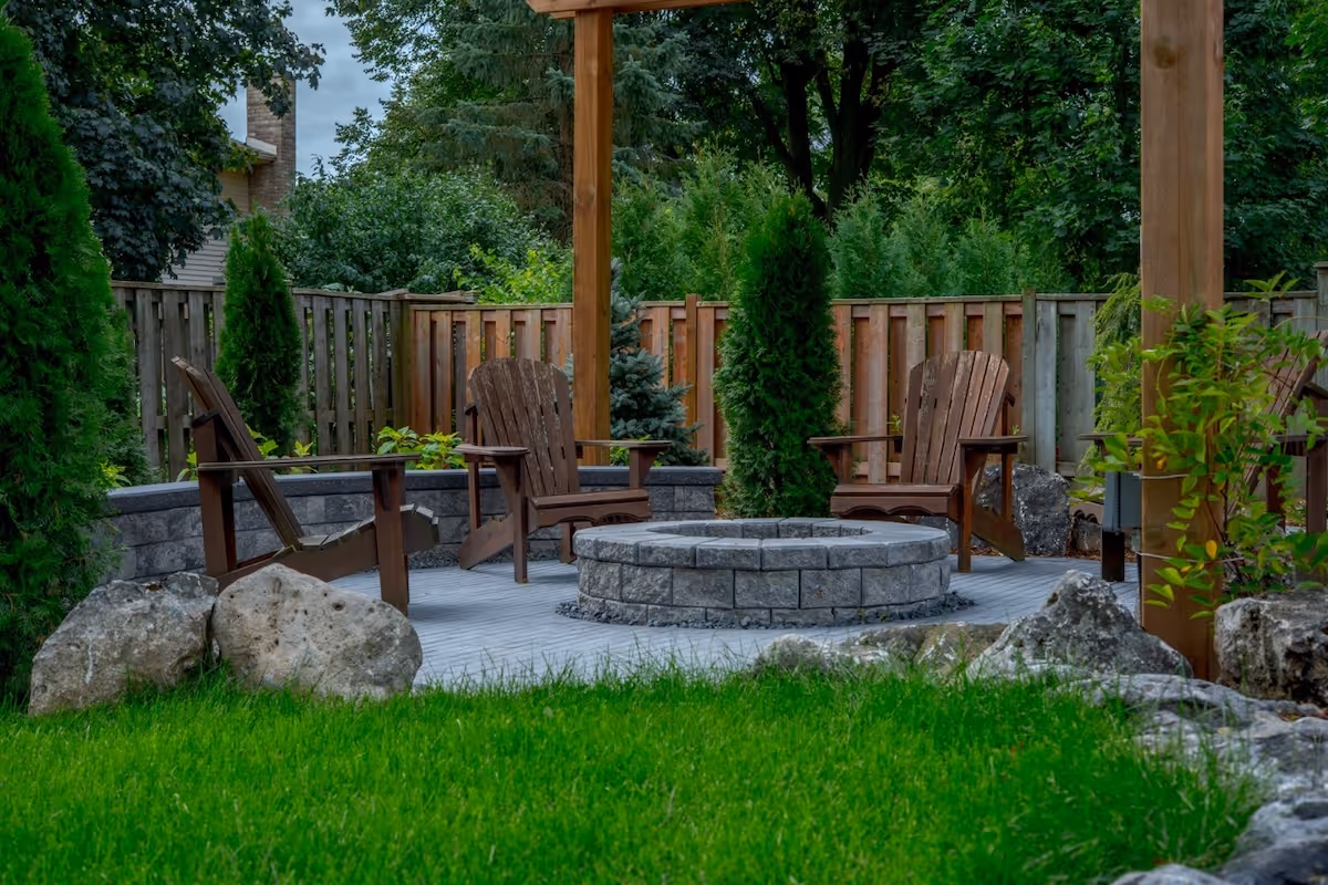 Backyard seating area with wooden chairs arranged around a stone fire pit, surrounded by greenery and a wooden fence.