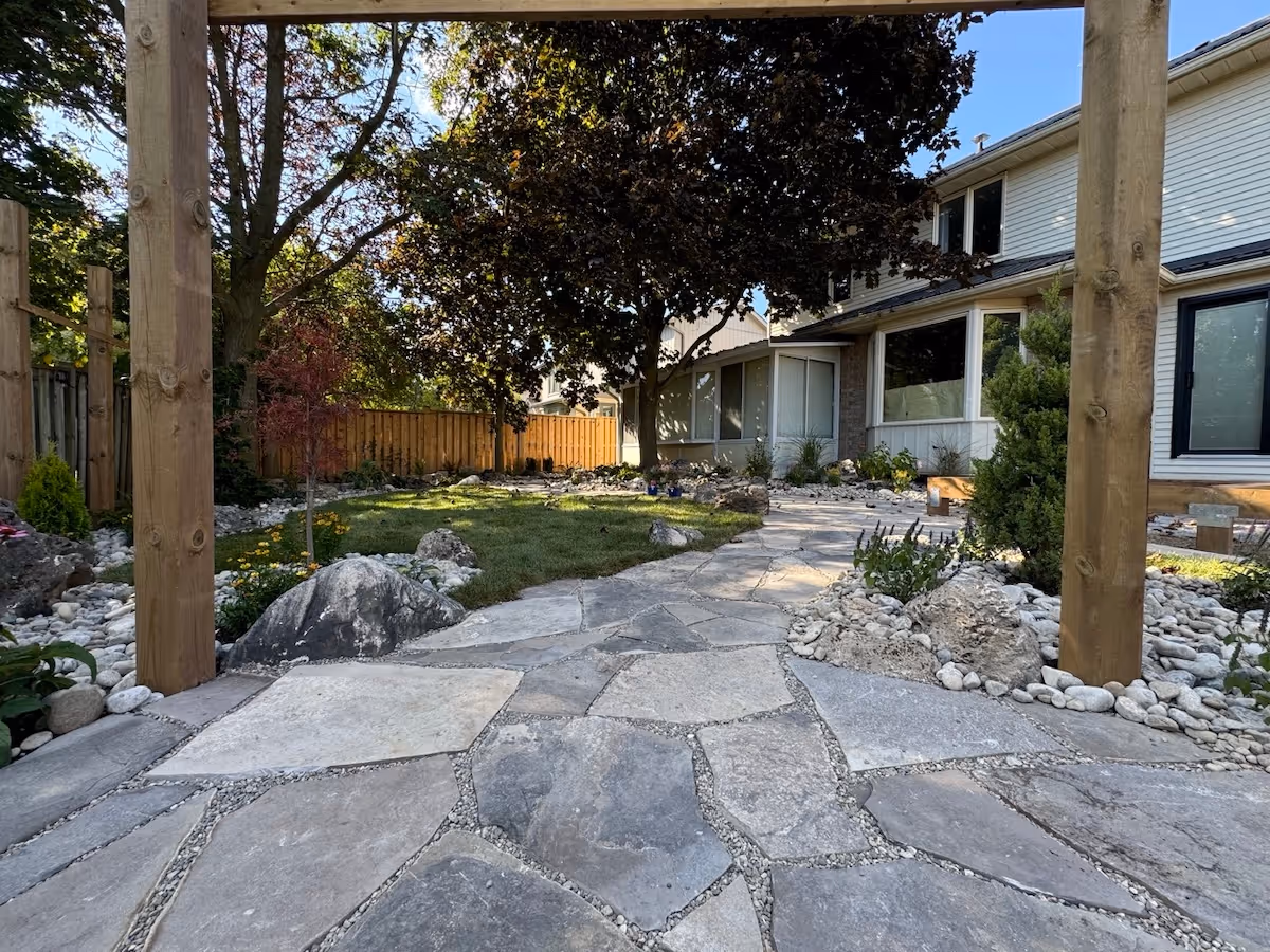 Stone pathway leading through a landscaped backyard with rocks, plants, and a large tree near a two-story house.
