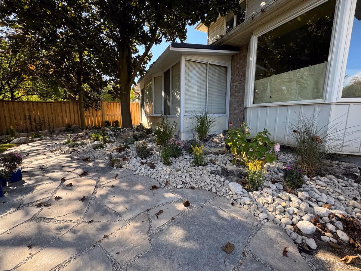 A stone-paved patio next to a house with a garden bed of white rocks, plants, and trees, bordered by a wooden fence.