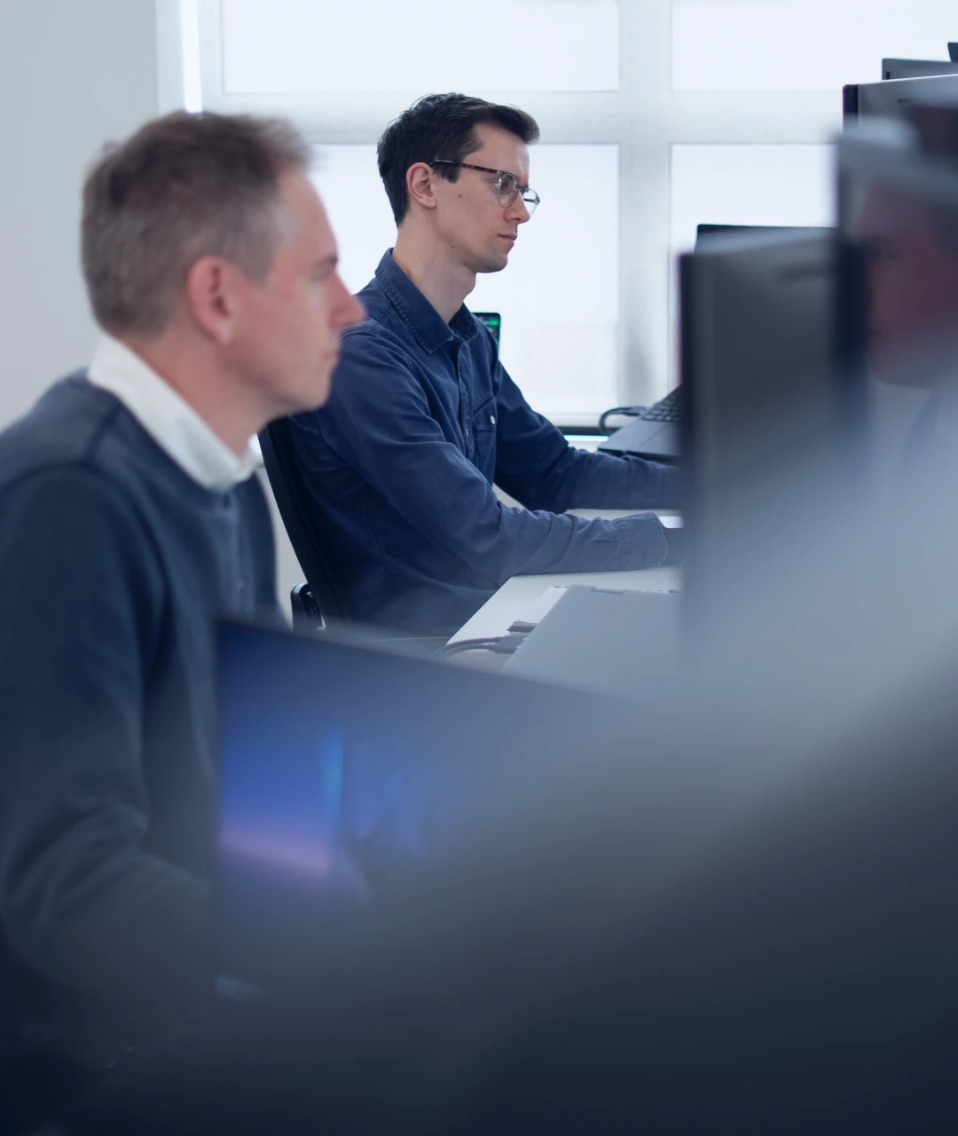 Two men seated in an office working on desktop computers near a large window.